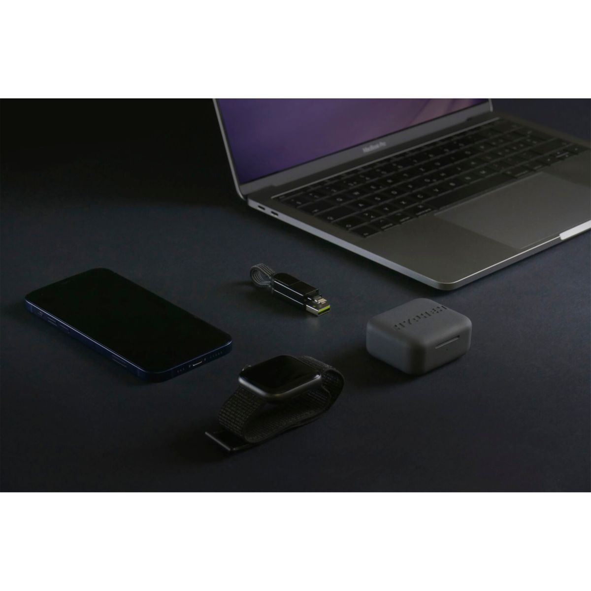 Un laptop, un telefono, un orologio, un cavo di ricarica e una custodia sono disposti su una superficie scura.