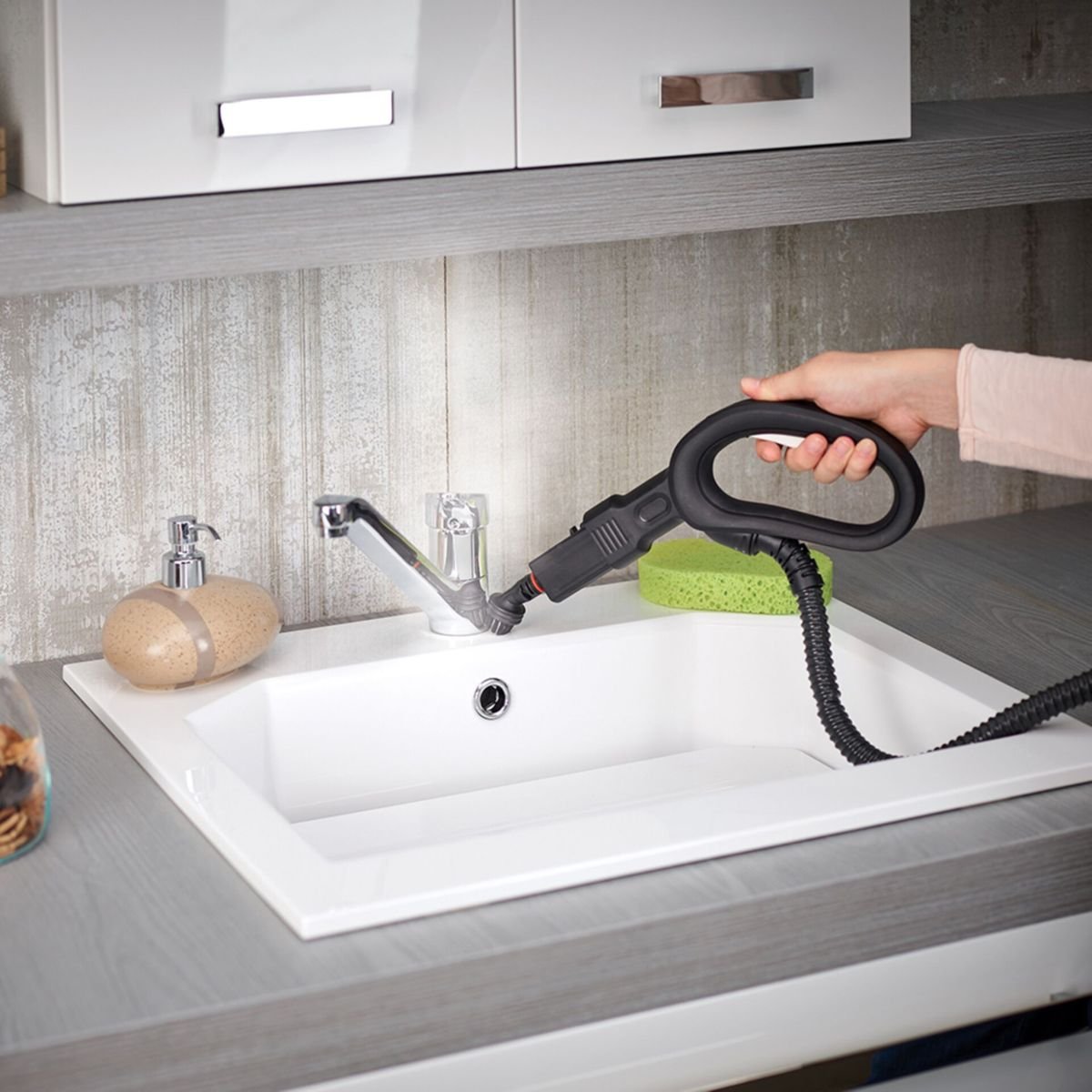 Osoba czyszcząca zlew za pomocą ręcznego parownika. Biały zlew, szary blat i szare szafki.