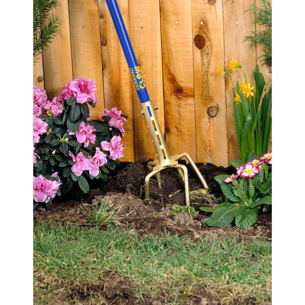 Een tuinklauwgereedschap in gebruik. Het staat in de grond bij roze bloemen en een houten hek.