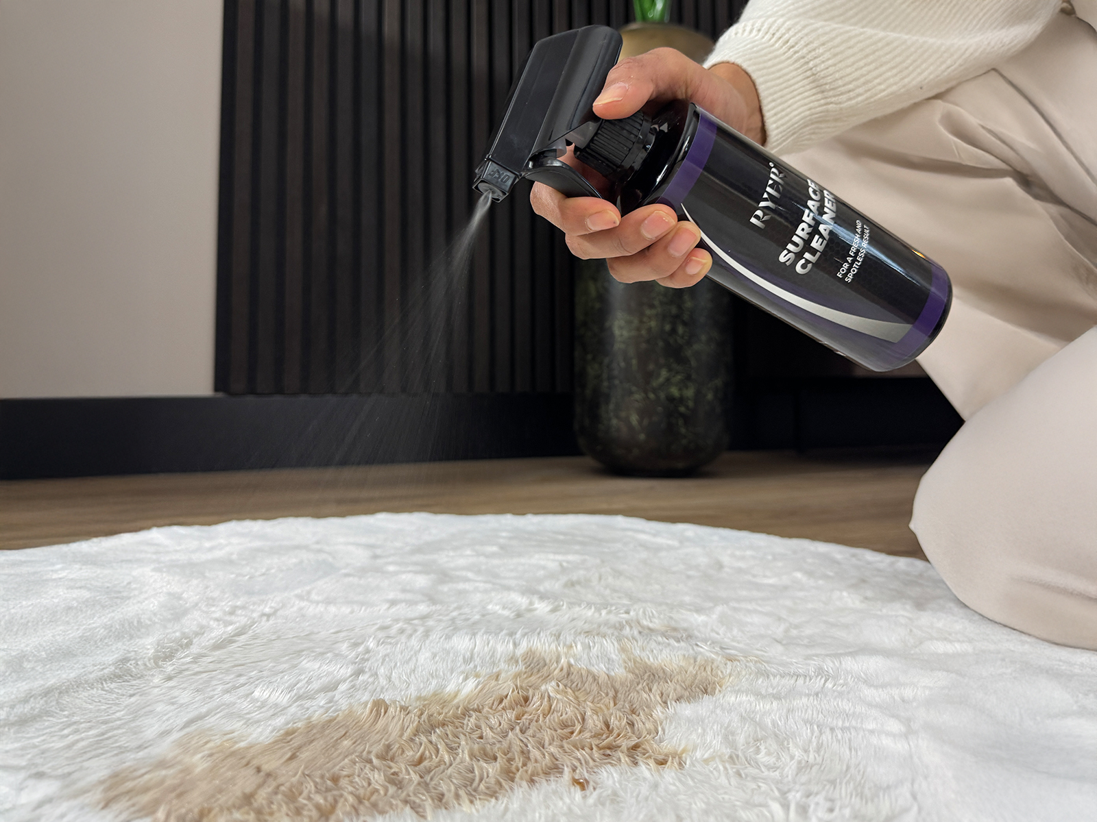 Een persoon spuit RYER Surface Cleaner op een wit tapijt met een bruine vlek.