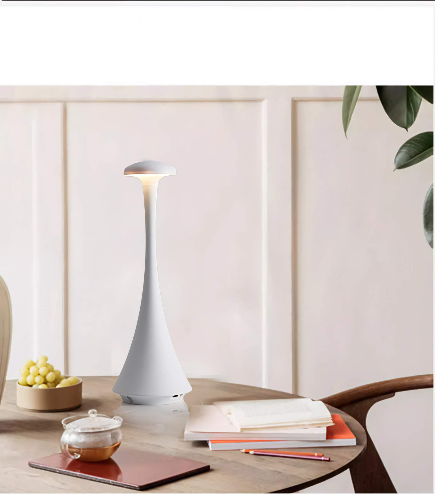 Weiße, moderne Tischlampe mit weißem Lampenschirm. Sie steht auf einem Holztisch mit Teeservice und Obstschale.