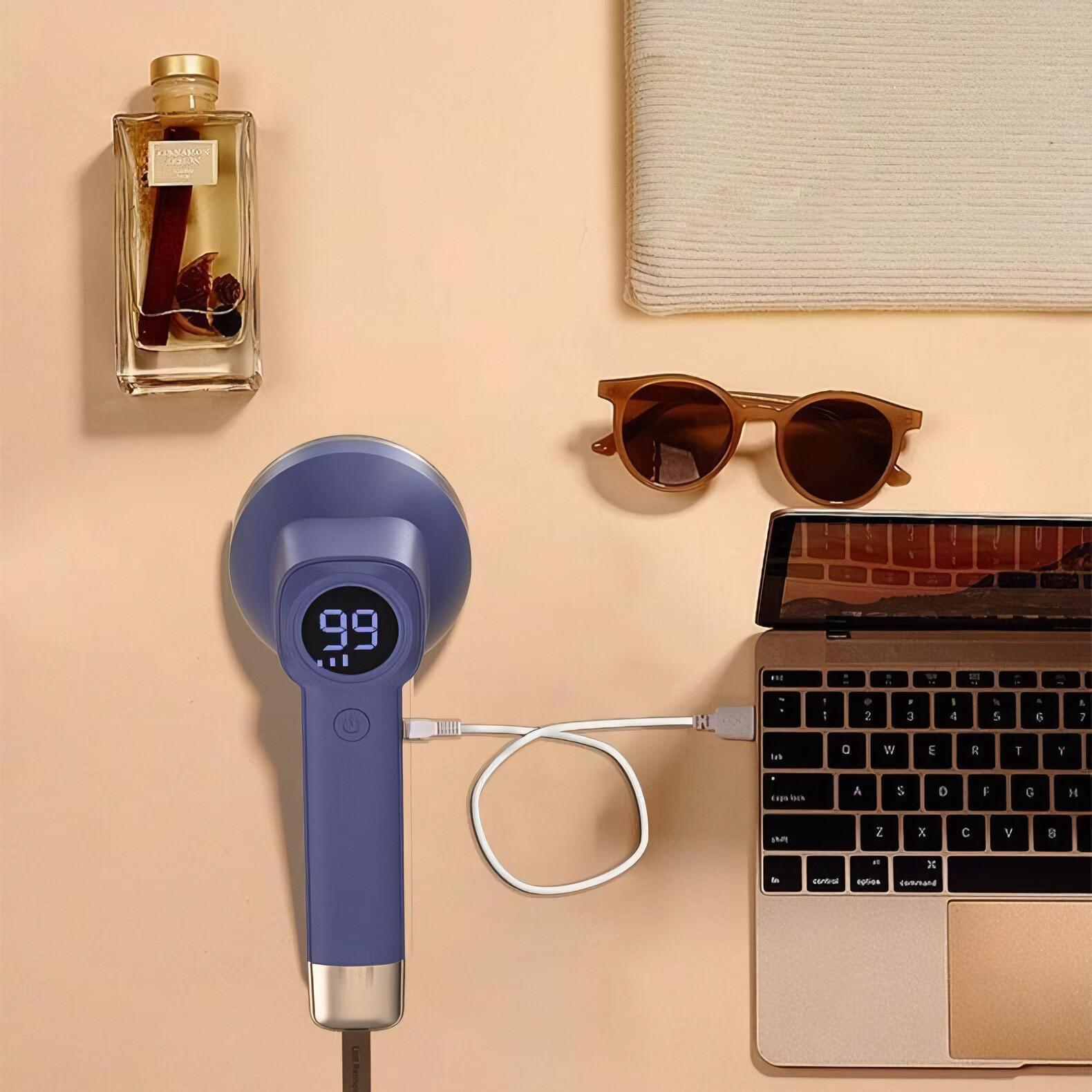 Ein blauer Stoffrasierer mit Bildschirm, neben einem Laptop, einer Sonnenbrille und einer Parfümflasche. Beige Hintergrund.