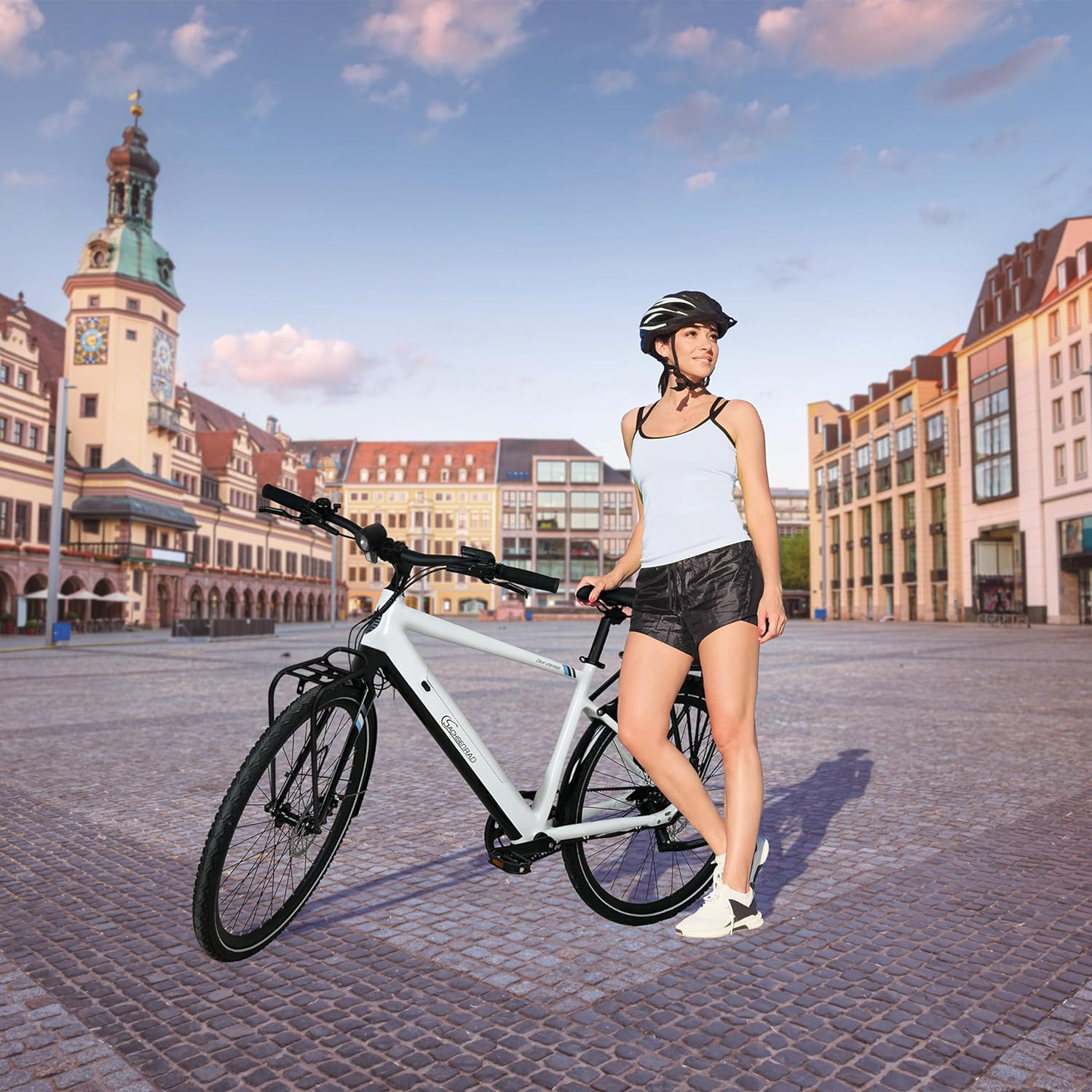 Eine Frau steht neben einem weißen Fahrrad mit schwarzem Gepäckträger auf einem Stadtplatz. Gebäude und blauer Himmel dahinter.