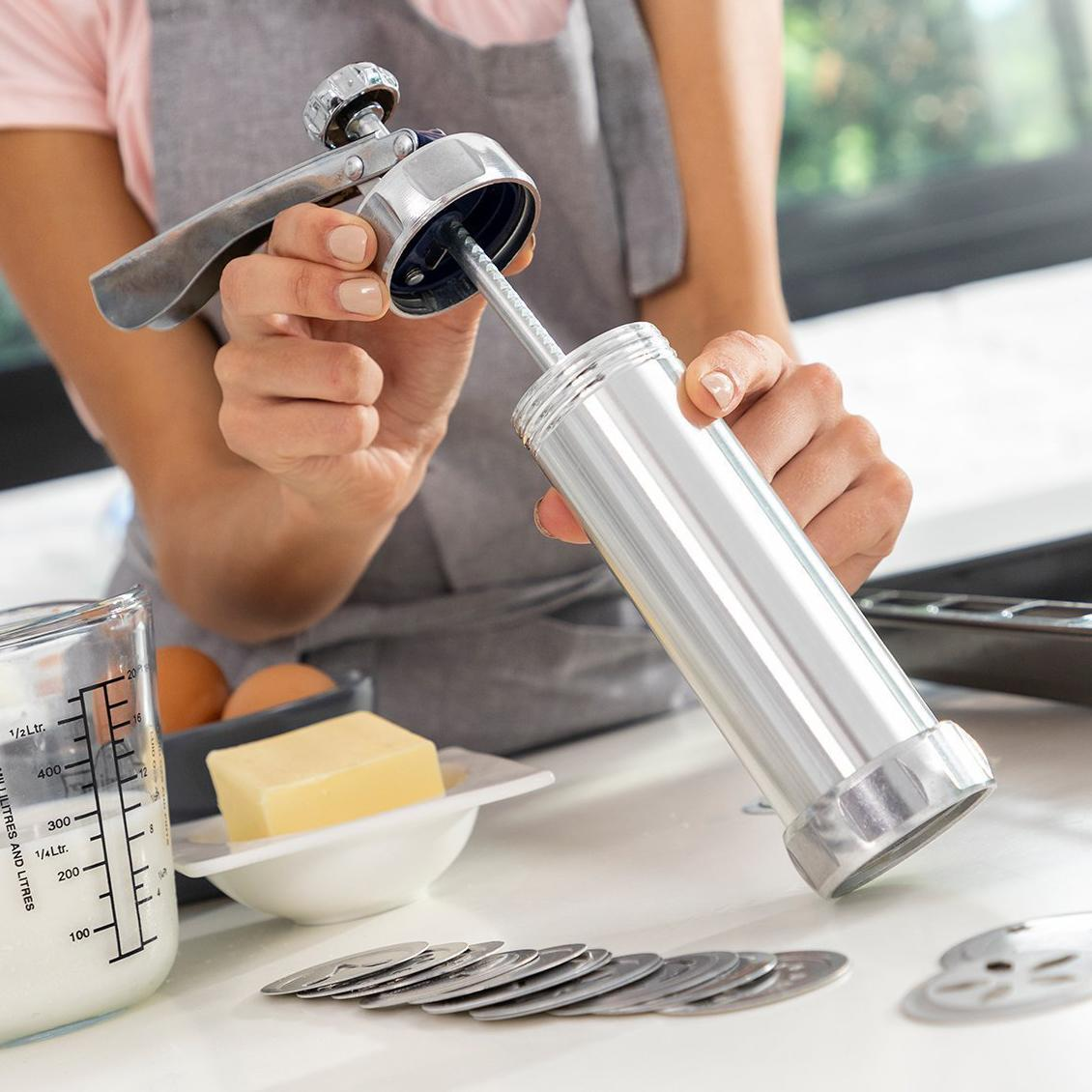 Mani che tengono una pressa per biscotti argentata con ingredienti e un misurino su un tavolo.