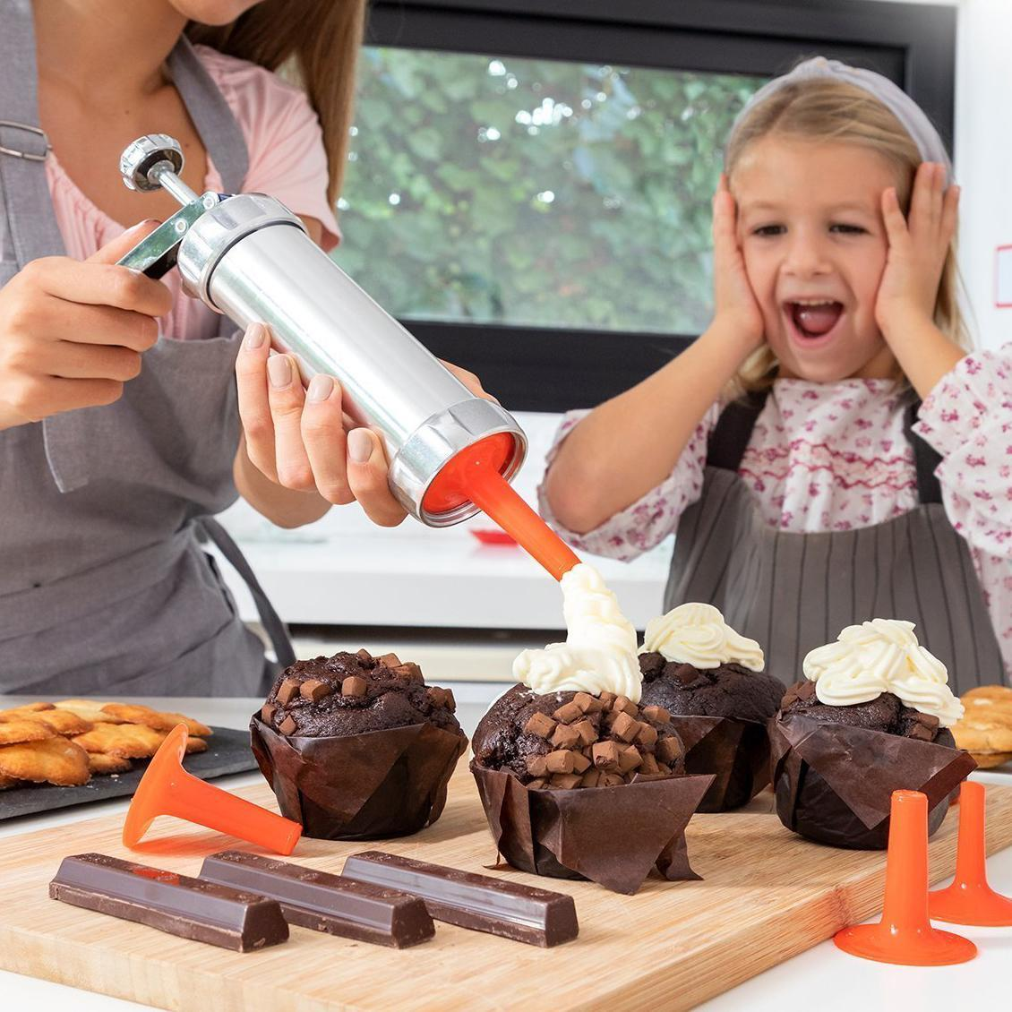 Una persona decora cupcake con crema bianca da uno strumento d'argento, con un bambino eccitato vicino.