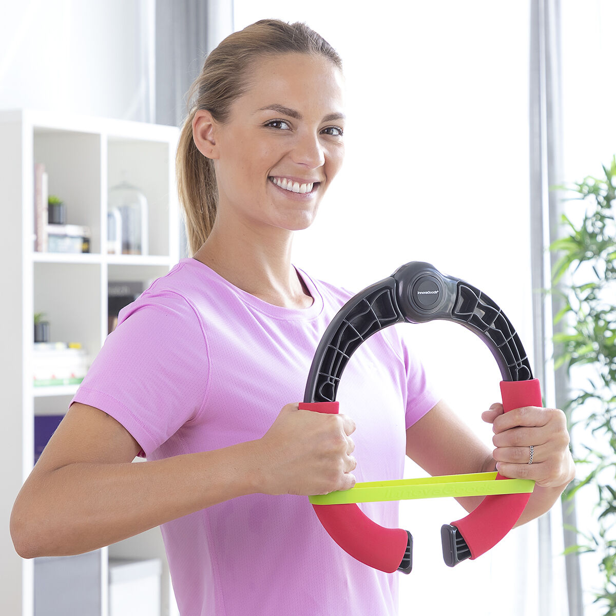 Lächelnde Frau hält einen Fitnessring. Sie trägt ein rosa Hemd. Hintergrund: Fenster und Regale.