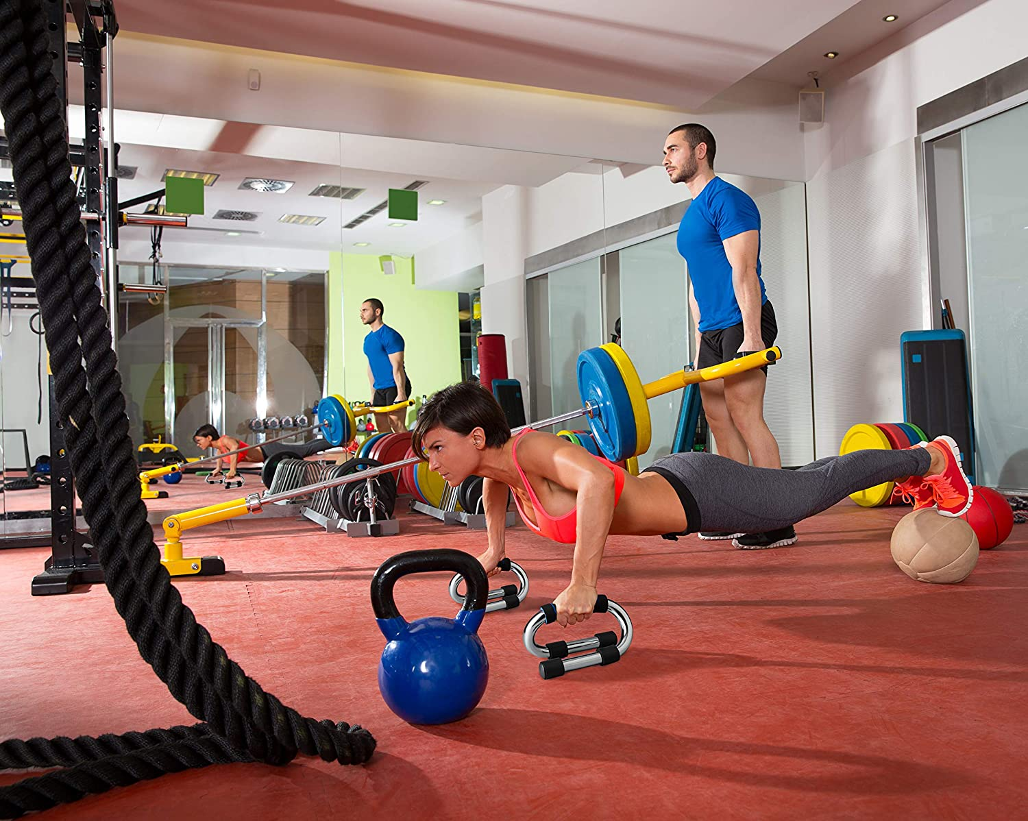 Frau macht Liegestütze mit Gewichten in einem Fitnessstudio. Mann hilft. Andere Leute trainieren im Hintergrund.
