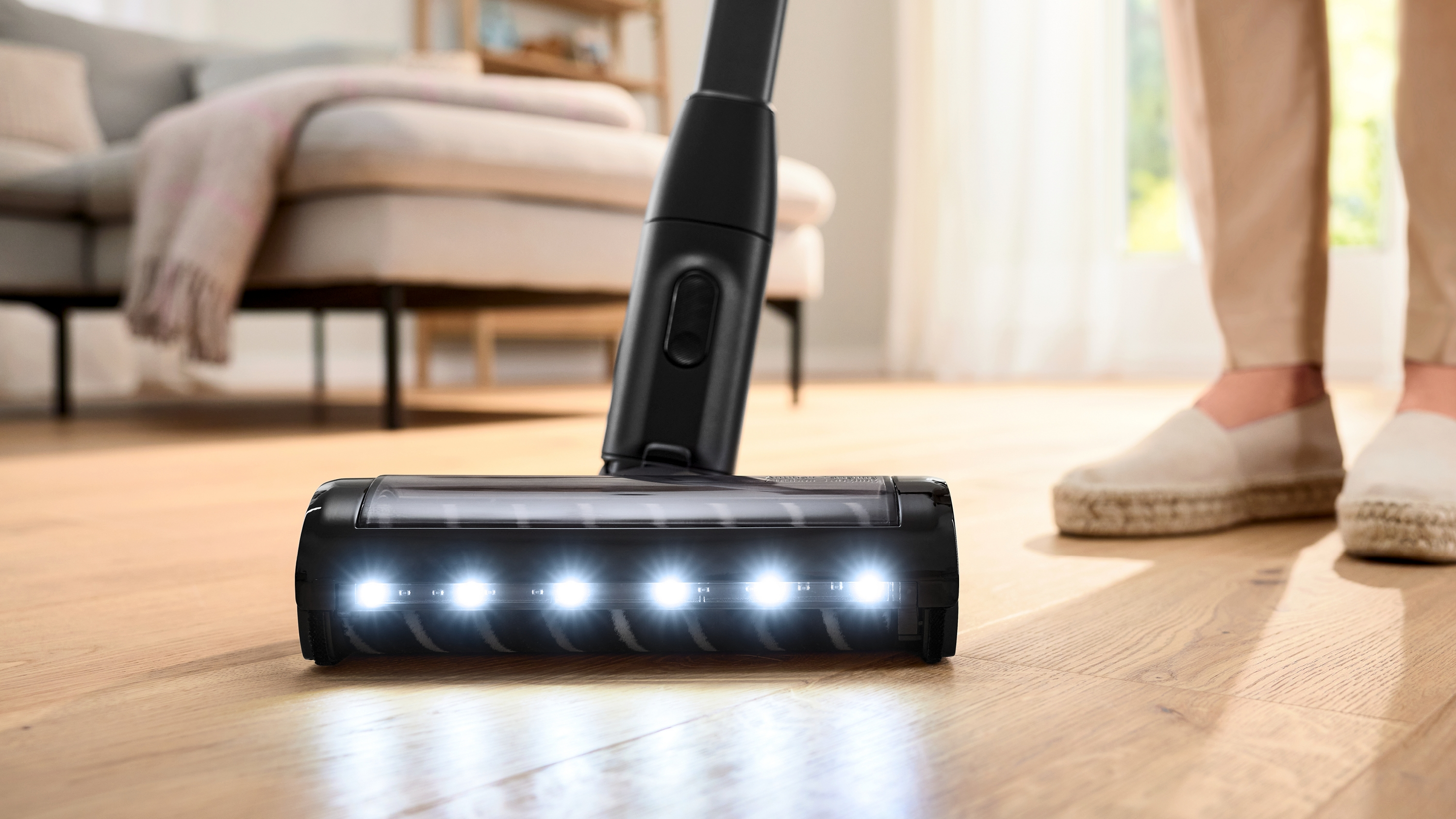 Un cabezal de aspiradora limpia un piso de madera. Tiene luces. Se ven las piernas y zapatos blancos de una persona.