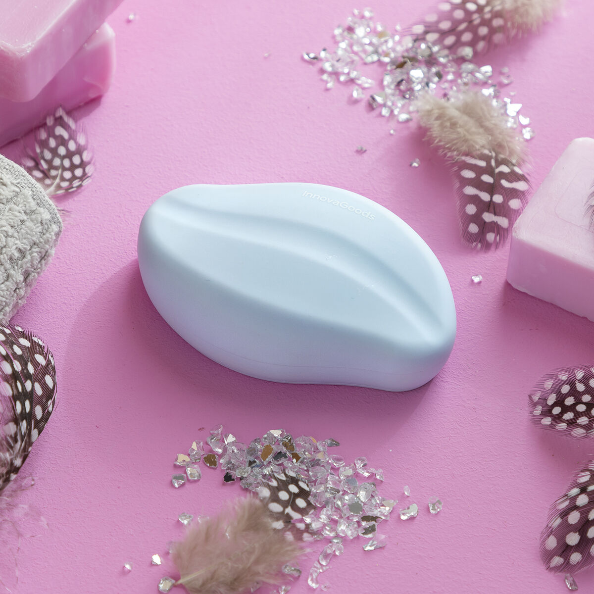 Un sapone blu ovale su sfondo rosa, circondato da piume, saponi e cristalli lucenti.