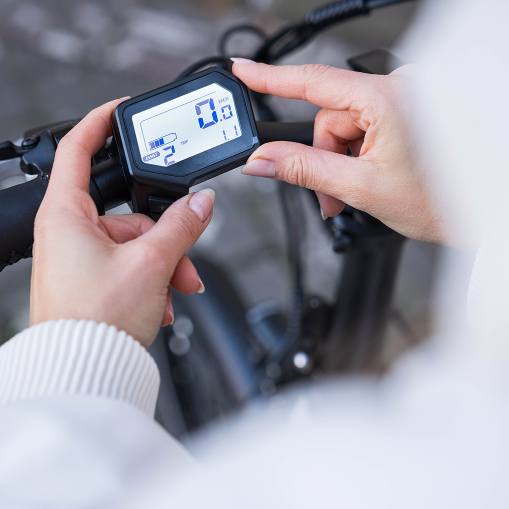 Mani che tengono il display di una e-bike che mostra velocità e informazioni sulla batteria. Sfondo sfocato.