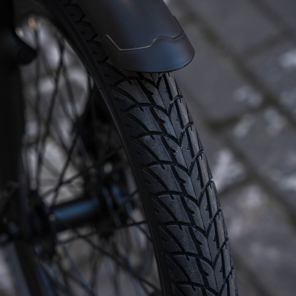 Primo piano di uno pneumatico nero per bicicletta con battistrada strutturato, su sfondo di mattoni grigi. Un parafango nero è sopra lo pneumatico.