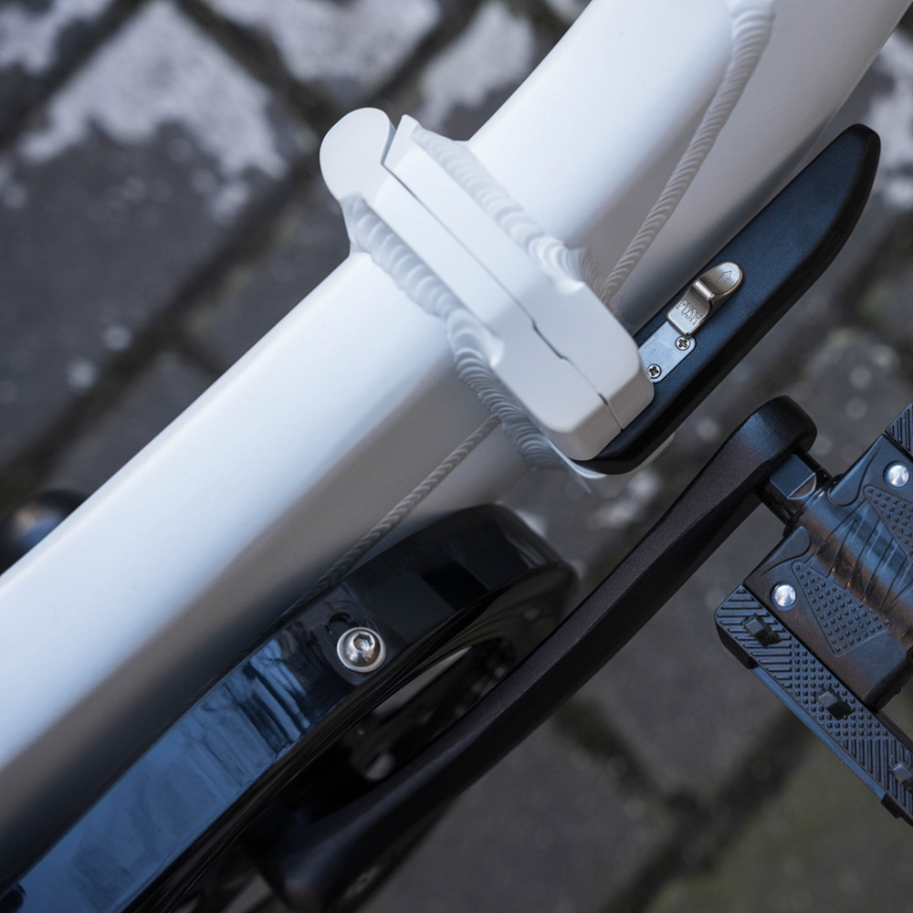 Primo piano di un telaio di bicicletta bianco, componenti neri e sfondo in mattoni grigi. L'attenzione è sulla serratura e sul pedale.