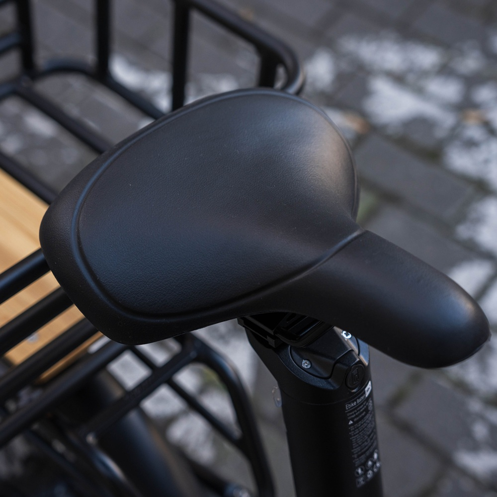 Primo piano di un sellino nero su una bici cargo. Lo sfondo è un ambiente urbano sfocato.