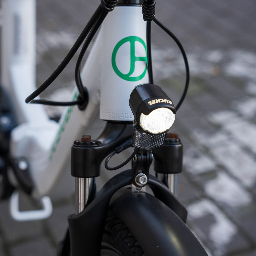 Primo piano del faro anteriore di una e-bike bianca. Il telaio della bici ha un logo verde. Sfondo sfocato.