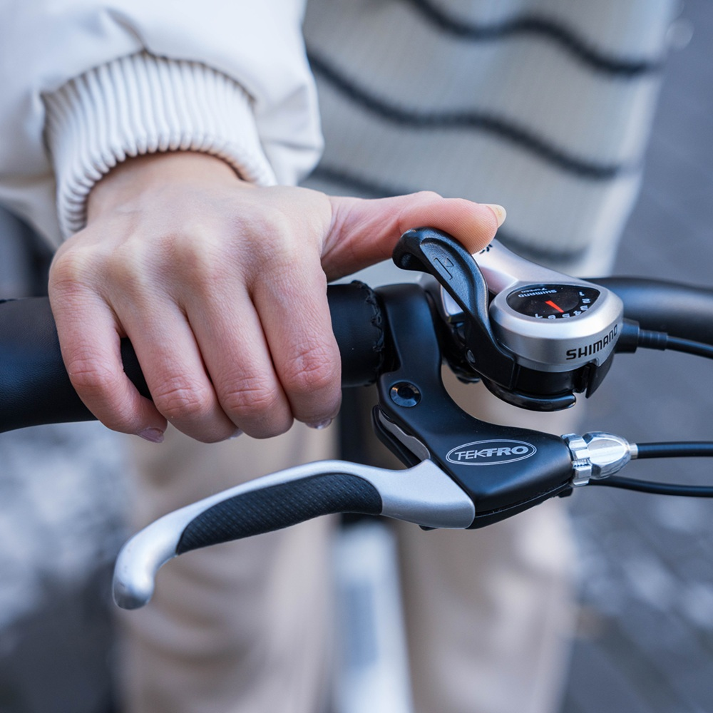 Primo piano di una mano che tiene il manubrio di una bici con indicatore marce Shimano e leva freno Tektro.