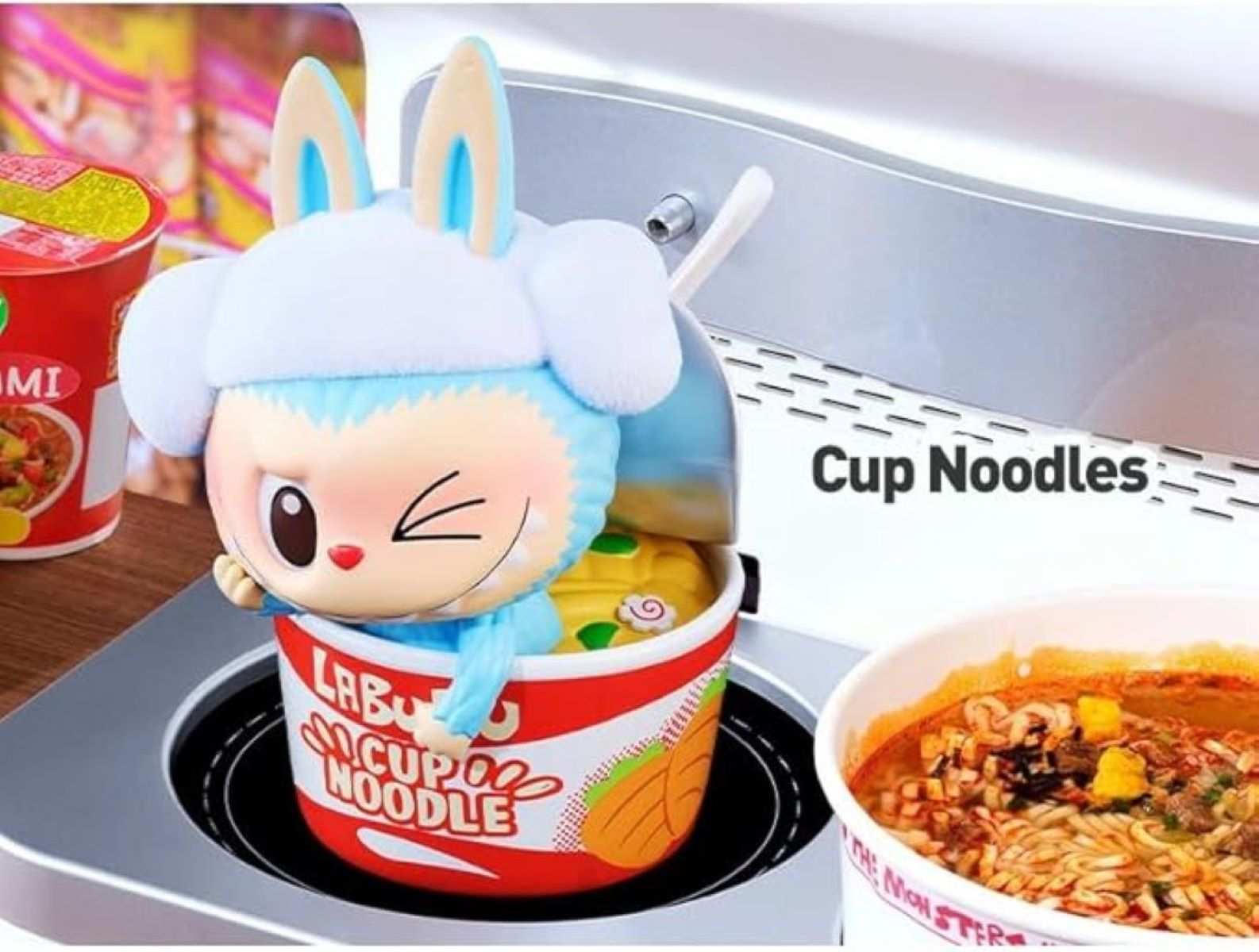 Un personaggio giocattolo in una tazza di noodles, con un cappello blu, che fa l'occhiolino accanto ad altri noodles.