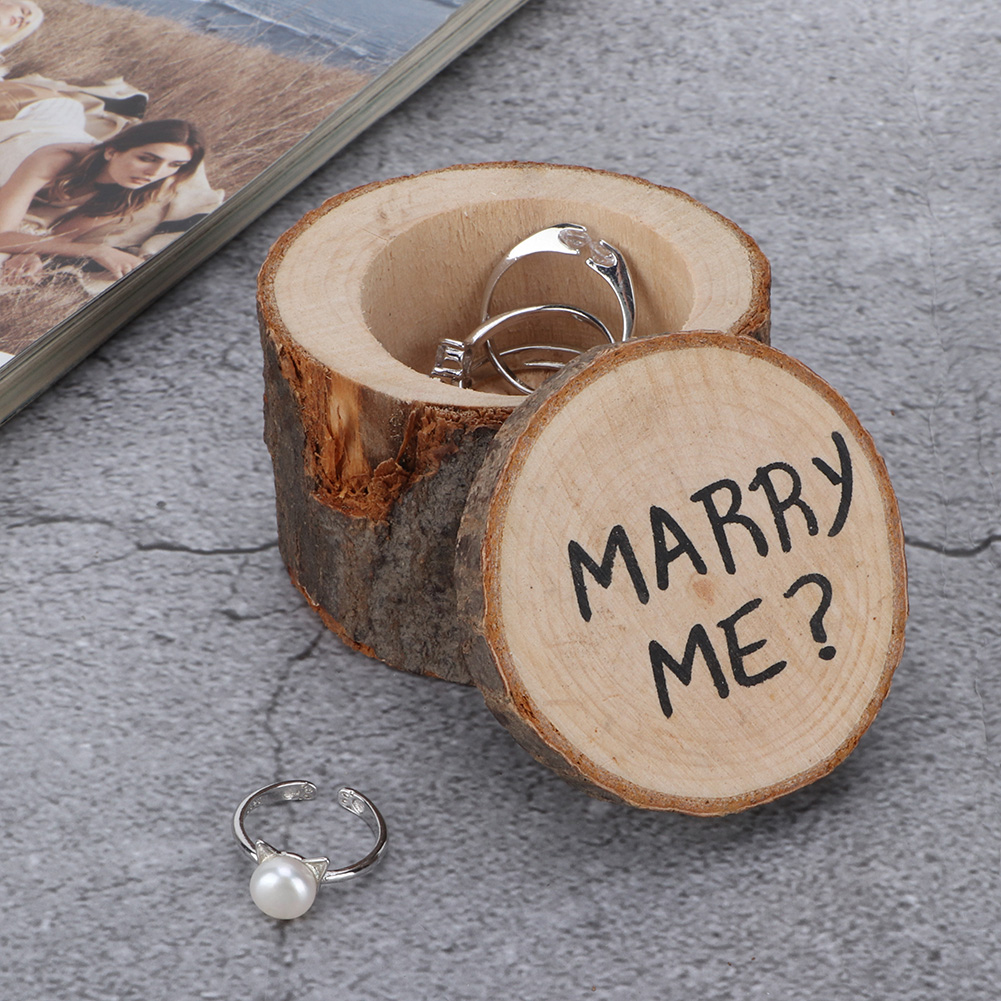 Holzringbox, offen mit Ringen darin, und Deckel mit der Aufschrift 'MARRY ME?'. Ringe sind darin. Box steht auf grauer Oberfläche.