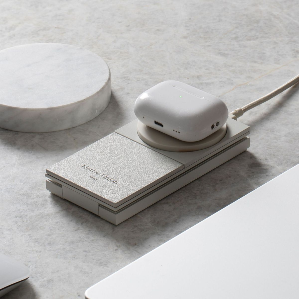 Weißes AirPods-Etui auf einem Ladepad. Das Pad befindet sich auf einer hellgrauen Oberfläche. Ein weiteres weißes Objekt im Hintergrund.