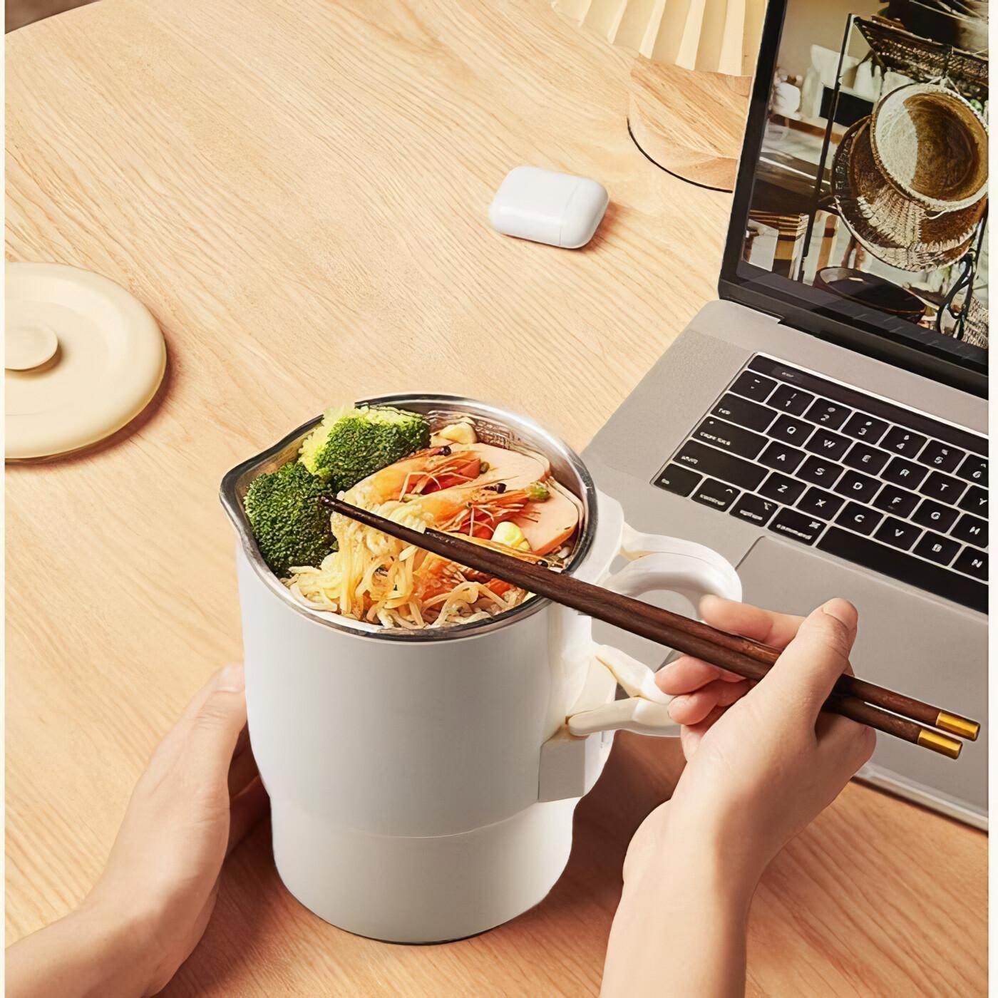 Person hält eine Schüssel mit Essen und Essstäbchen in der Nähe eines Laptops. Weiße Tasse, Holztisch.
