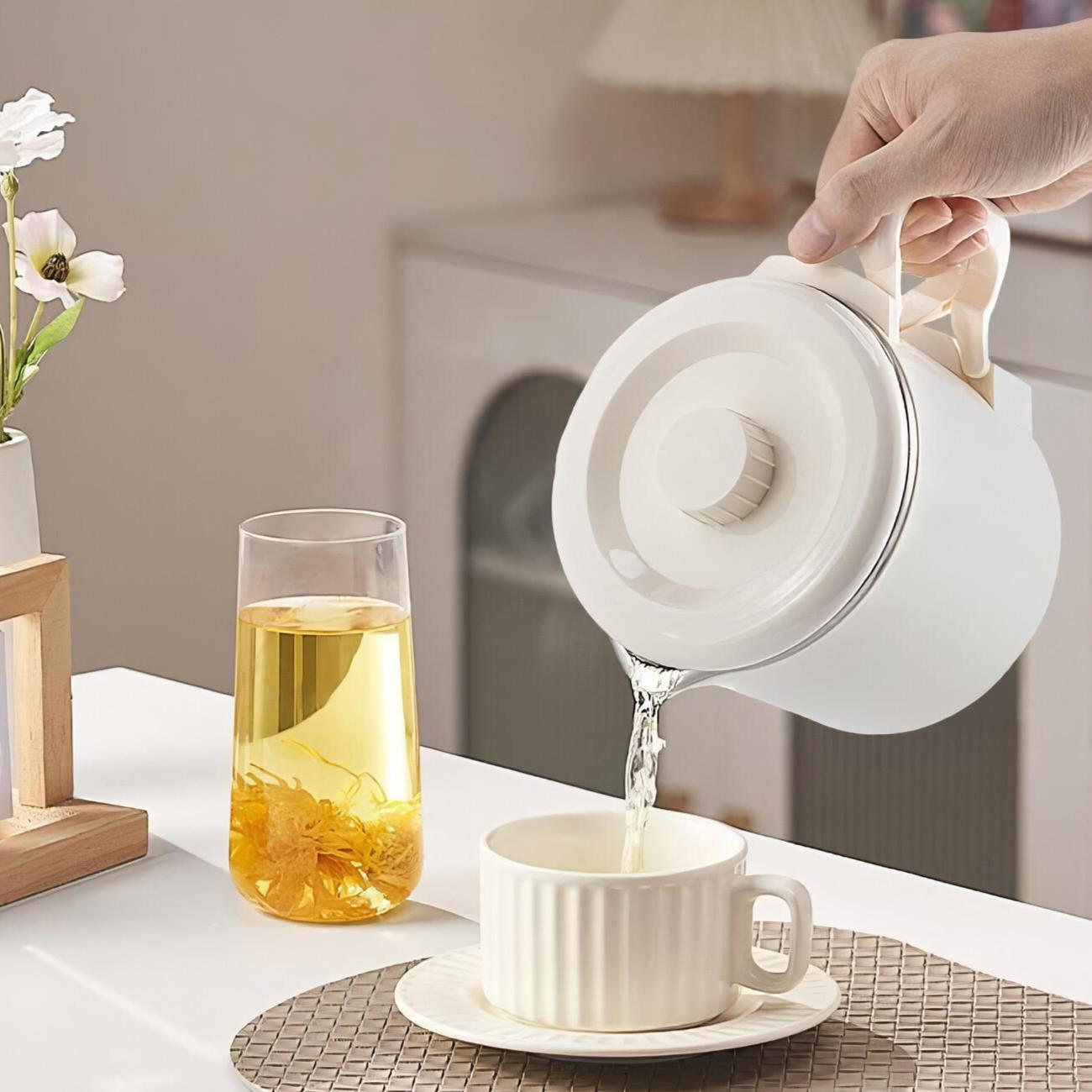 Eine Person gießt Wasser aus einem weißen Wasserkocher in eine weiße Tasse. Links ein Glas mit gelber Flüssigkeit.