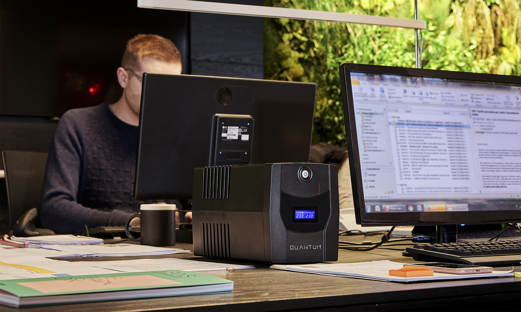 Un uomo che lavora a una scrivania con un UPS, monitor di computer e una tazza.