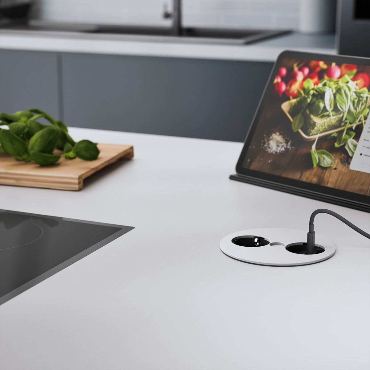 Un piano cucina con basilico, un tablet che mostra cibo e una presa di corrente. A sinistra un tagliere di legno.