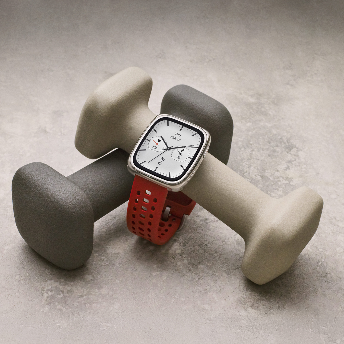 Smartwatch z czerwonym paskiem spoczywa na dwóch szarych i beżowych hantlach na jasnoszarej powierzchni.