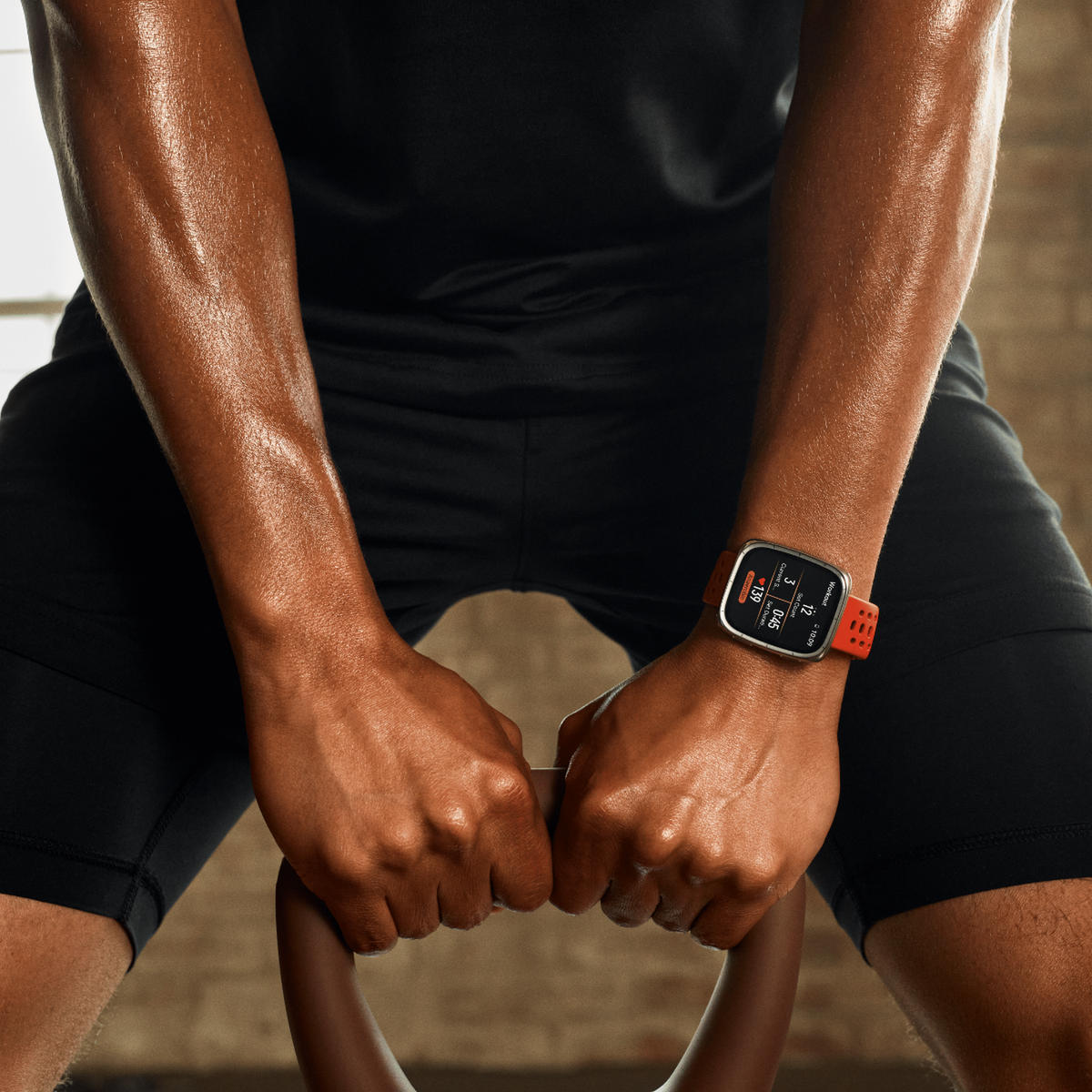 Osoba trzymająca kettlebell, nosząca smartwatch z czerwonym paskiem, na tle siłowni.
