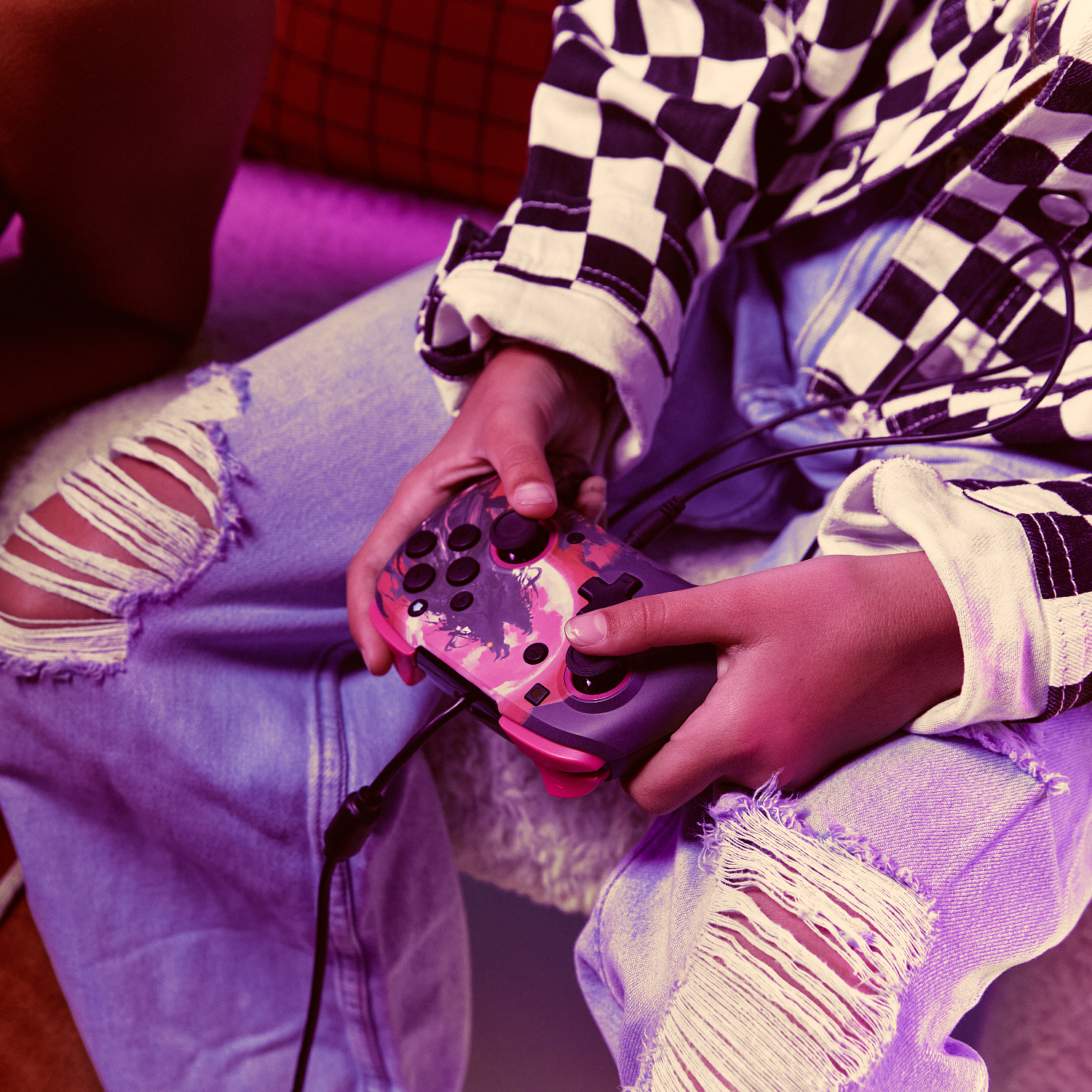 Osoba trzyma różowo-czarny kontroler. Ma na sobie podarte dżinsy i koszulę w kratę.