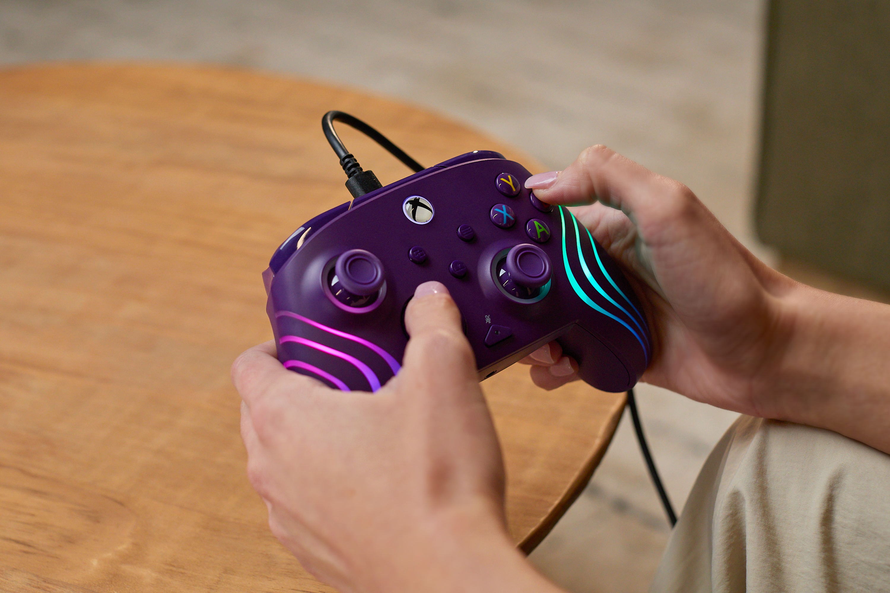 Dłonie trzymające fioletowy kontroler Xbox z tęczowymi światłami na drewnianym stole.