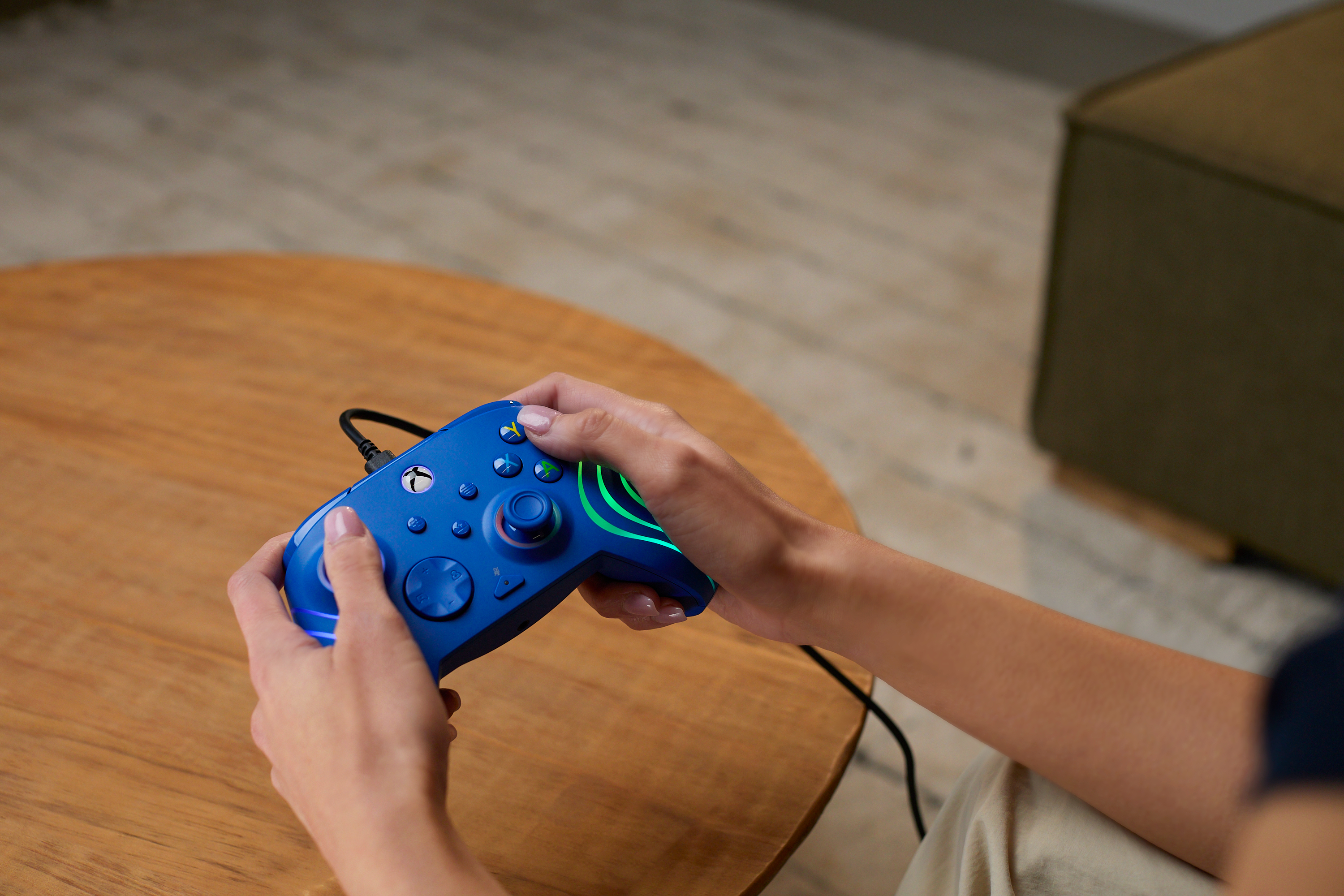 Ręce trzymające niebieski kontroler Xbox. Kontroler jest blisko drewnianego stołu.