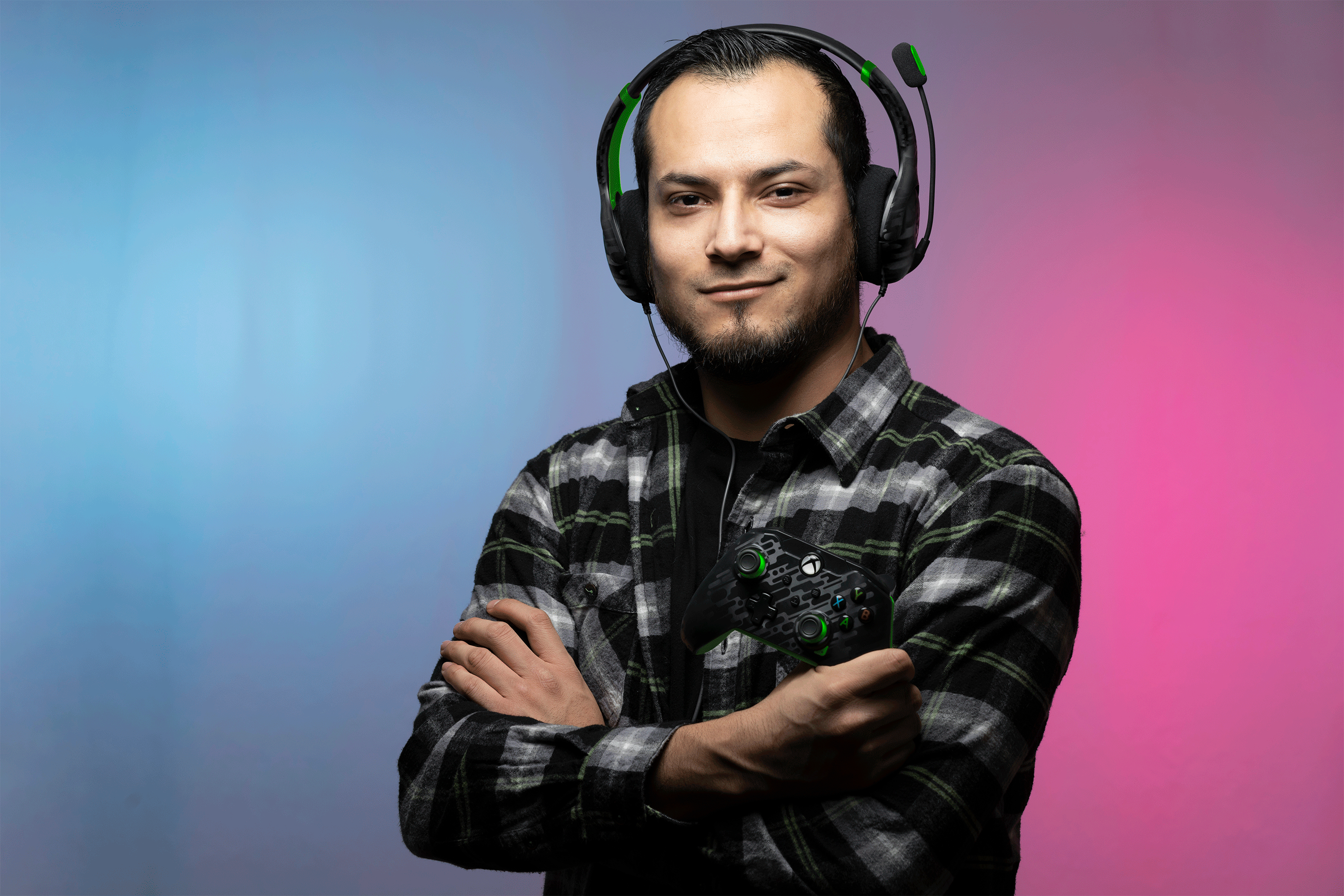 Un uomo con un controller Xbox e un auricolare. Indossa una camicia a quadri e guarda la fotocamera.