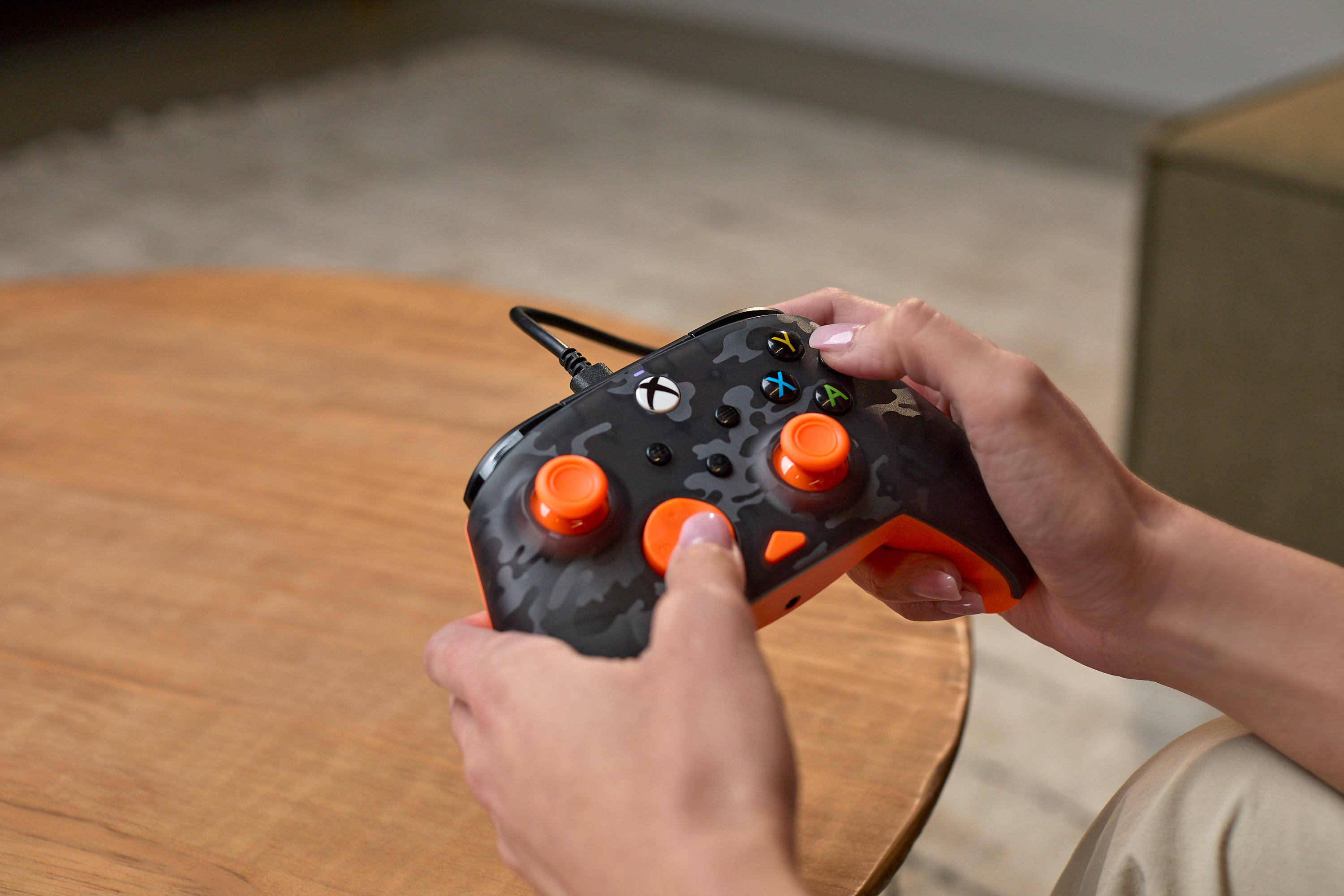 Osoba trzymająca kontroler Xbox z kamuflażem. Kontroler ma pomarańczowe akcenty.