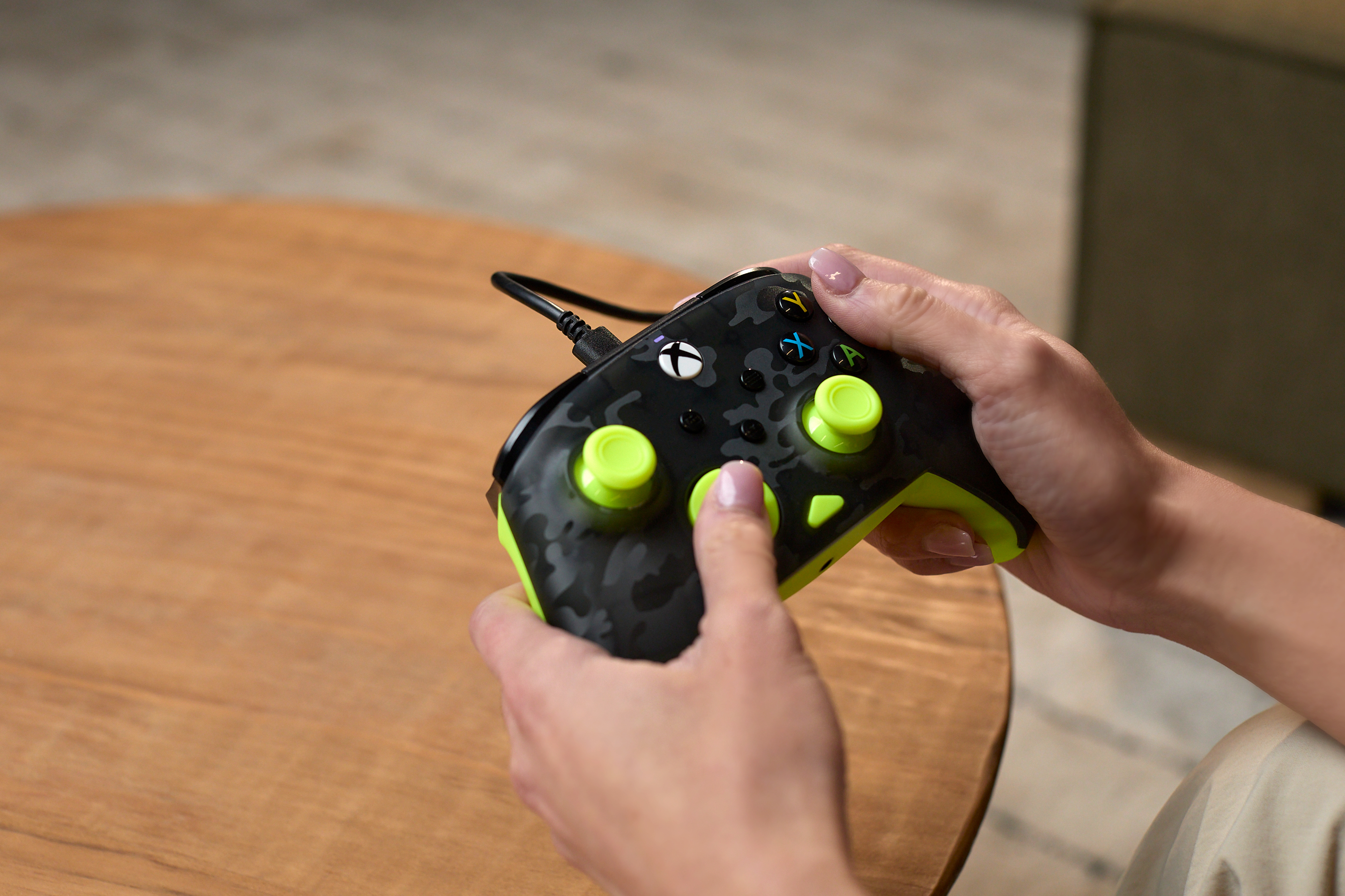Ręce osoby trzymającej kontroler Xbox w kamuflażu na drewnianym stole.