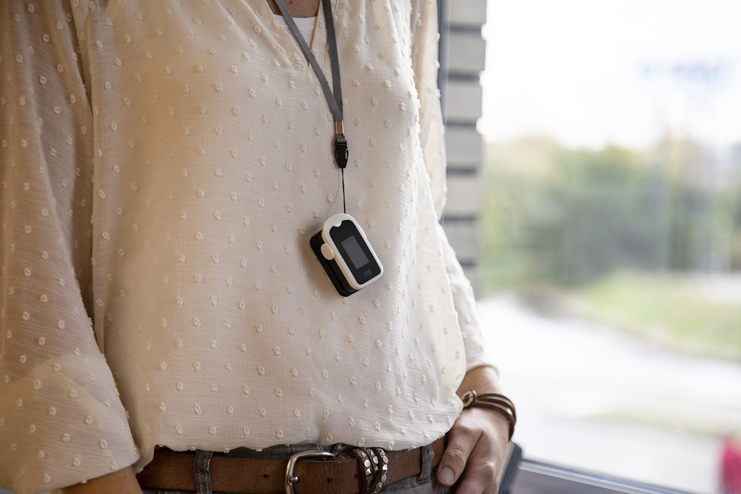 Eine Person trägt eine weiße Bluse mit einem Pulsoximeter, das an einem Lanyard hängt. Im Hintergrund sind ein Fenster und verschwommenes Grün.