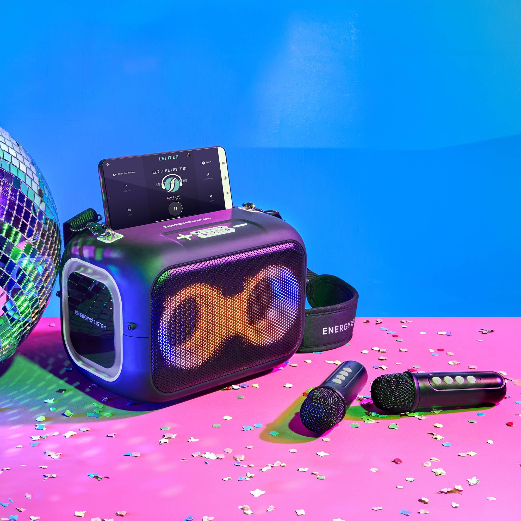 Un'impostazione di altoparlanti per karaoke neri con uno smartphone, due microfoni e una palla da discoteca. La scena è vibrante.