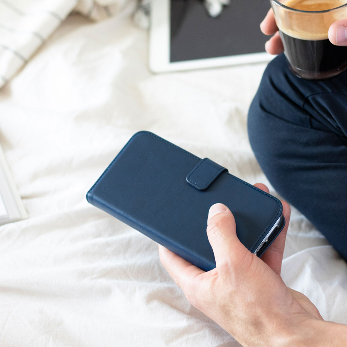 De handen van een persoon die een donkerblauwe telefoonhoes en een koffiekopje vasthoudt, met een tablet op de achtergrond.