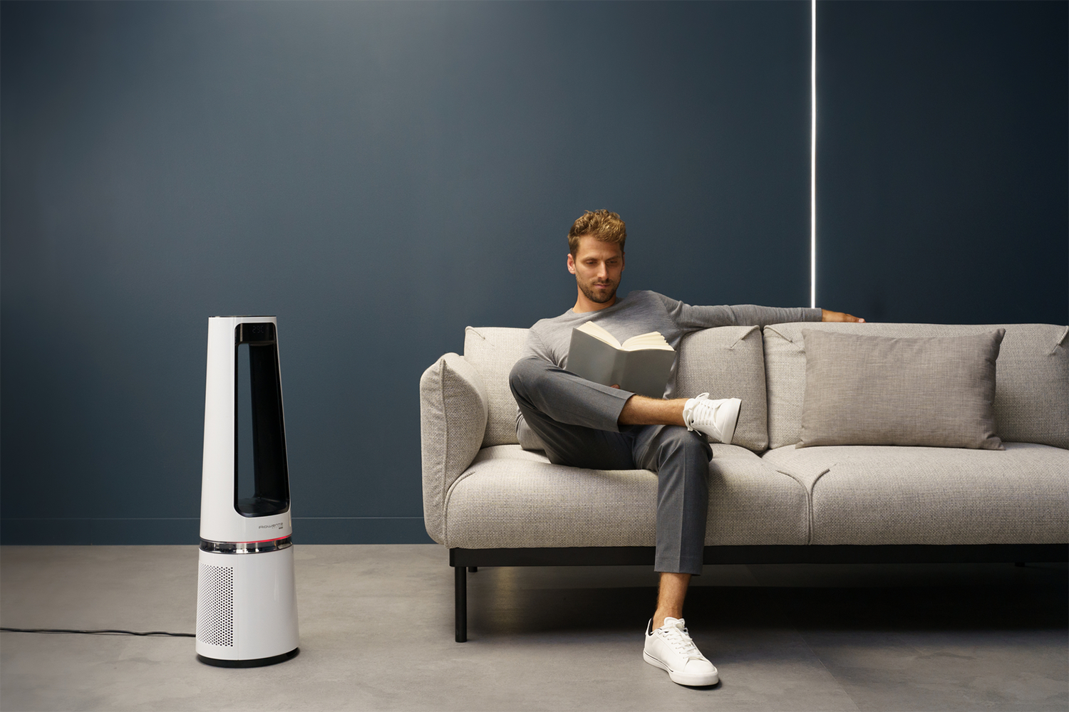 A man sits reading a book on a couch, next to an air purifier. The room has a dark blue wall and white trim.
