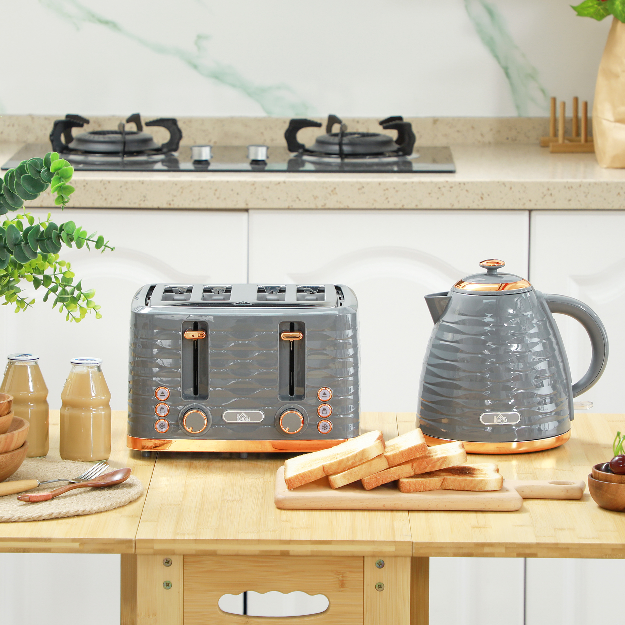 Ein grauer Toaster und ein Wasserkocher auf einem Holztisch mit Toastscheiben. Links ist eine grüne Pflanze.