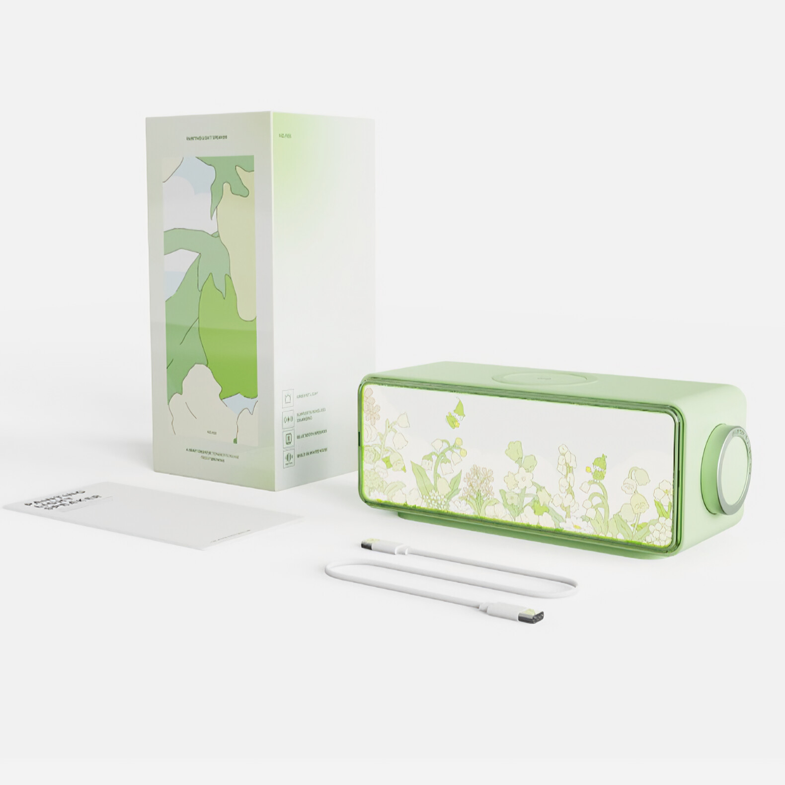 Ein Lautsprecher und eine Box mit floralem Muster. Die Box hat einen grünen Farbverlauf. Weißer Hintergrund und zwei Ladekabel.