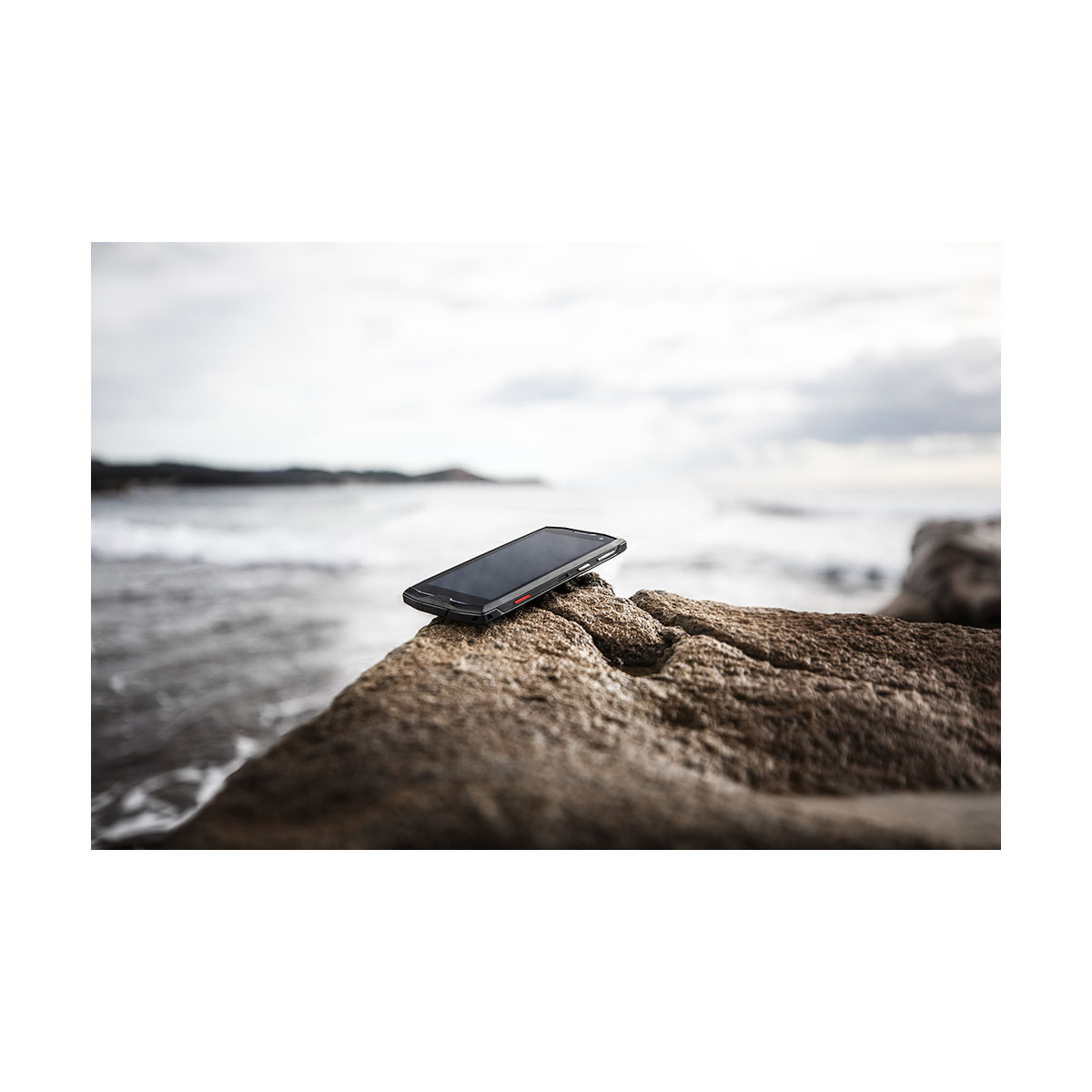 Un smartphone noir repose sur un rocher près de la mer. La mer est en arrière-plan avec un ciel nuageux.