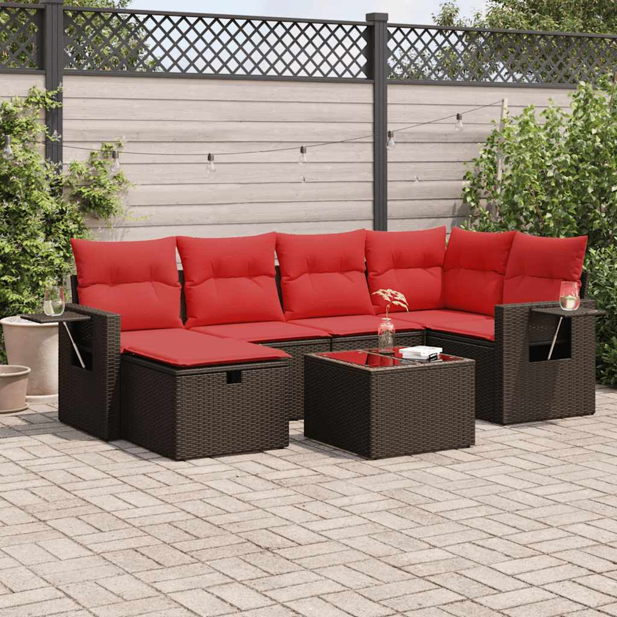 Un sofá seccional de mimbre con cojines rojos y una mesa con tapa de vidrio se encuentra en un patio de ladrillo en un jardín.