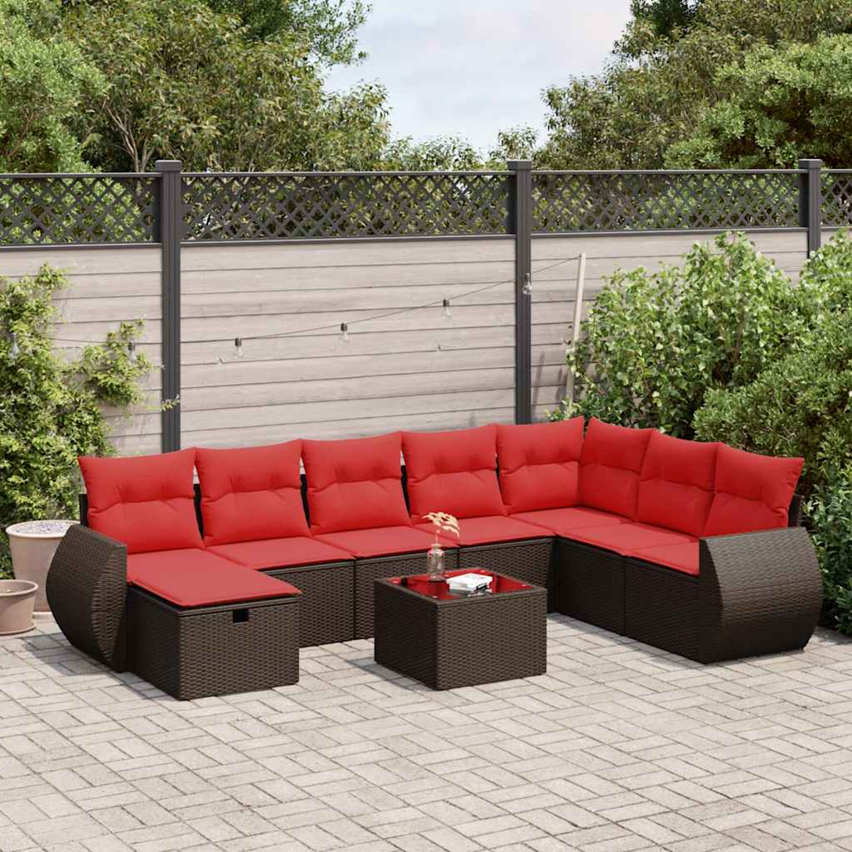 Eine geflochtene Eckcouch mit roten Kissen und einem Tisch mit Glasplatte steht auf einer Backsteinterrasse in einem Garten.