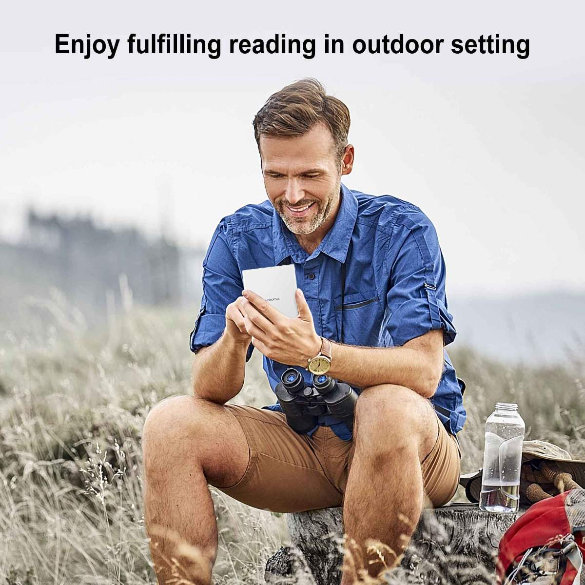 Mann liest E-Reader im Freien, lächelt, mit Fernglas und Wasserflasche.