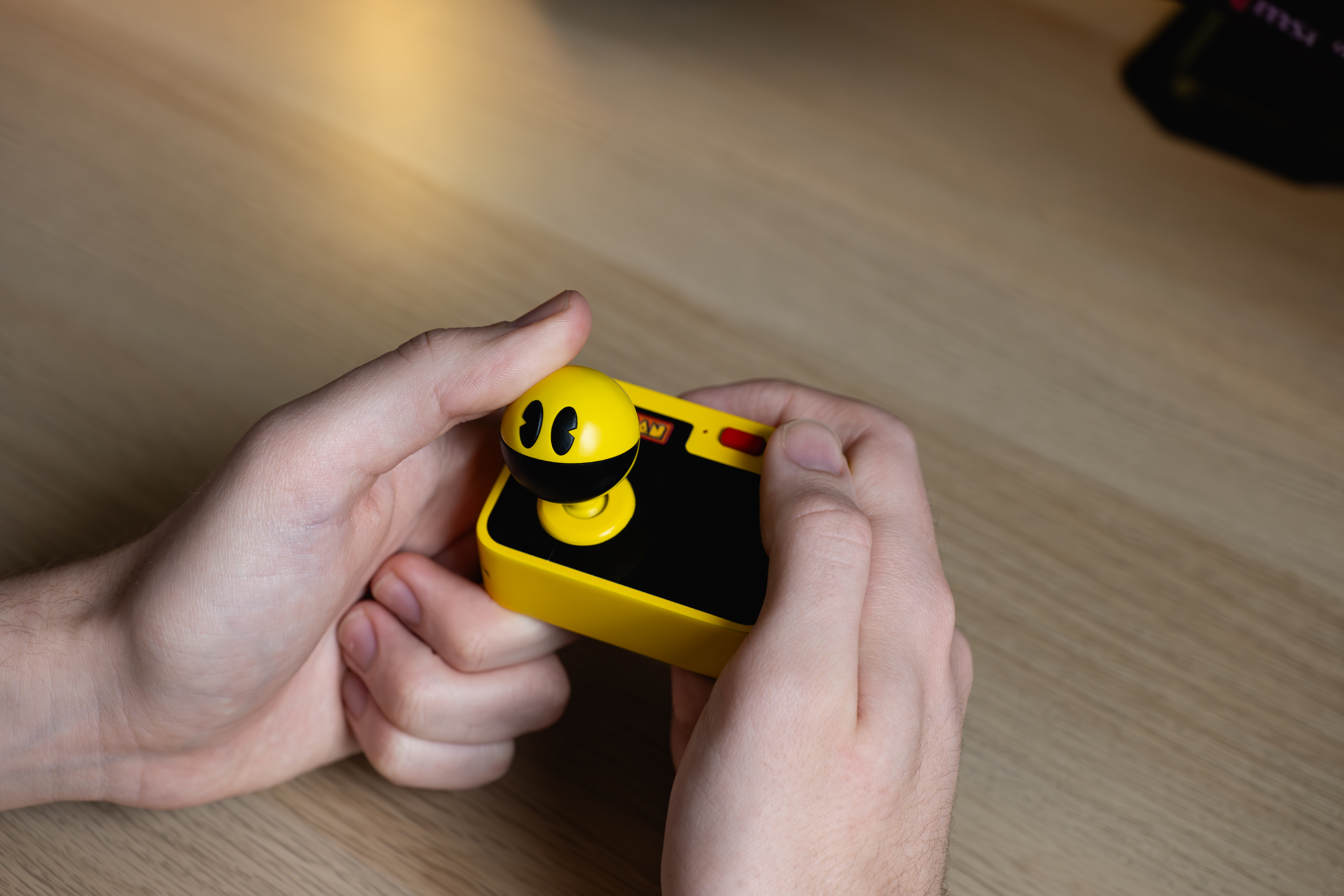 Handen die een gele Pac-Man joystick-controller vasthouden. Een houten oppervlak op de achtergrond.