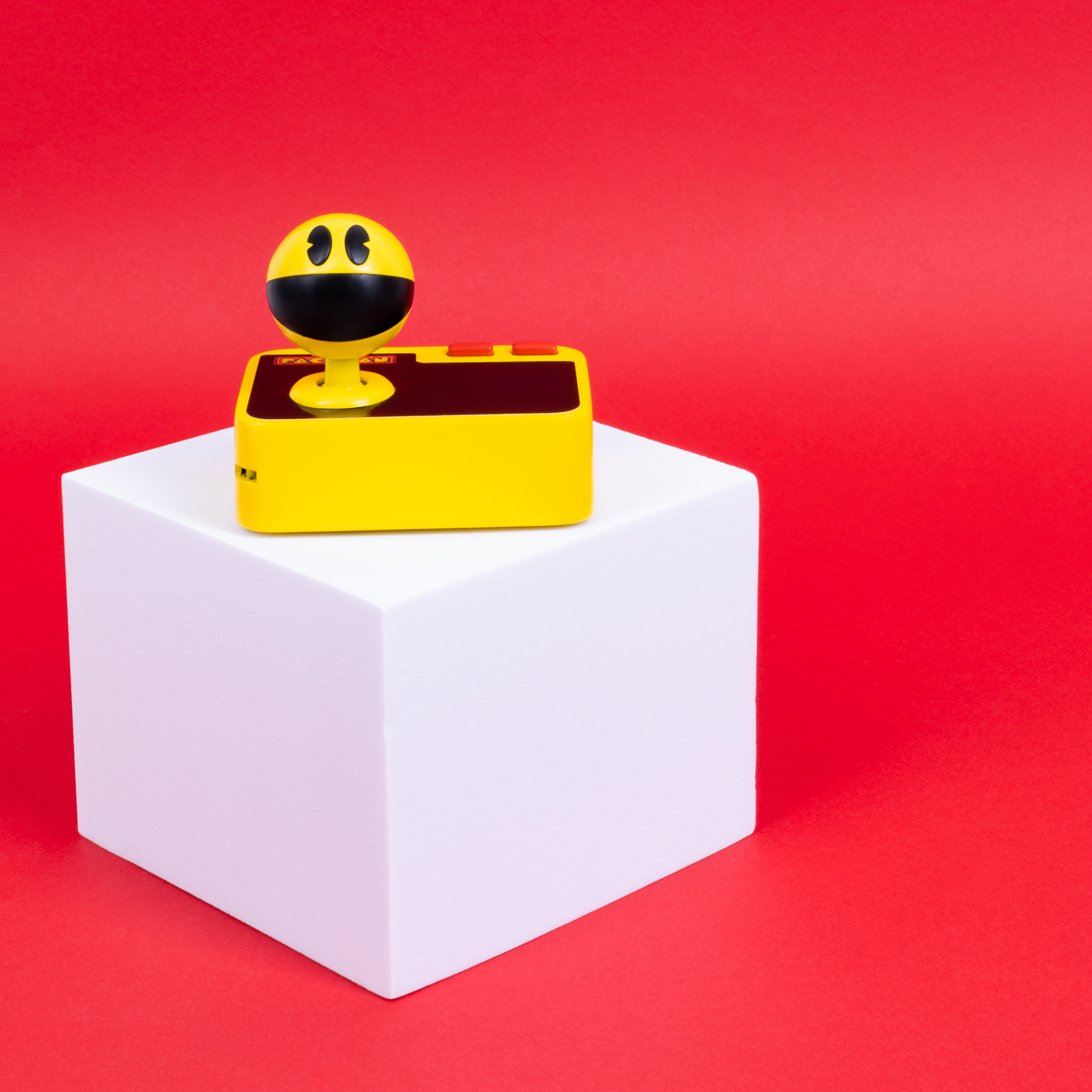 Gele Pac-Man-controller op een witte kubus tegen een rode achtergrond. Zwarte details op de controller.