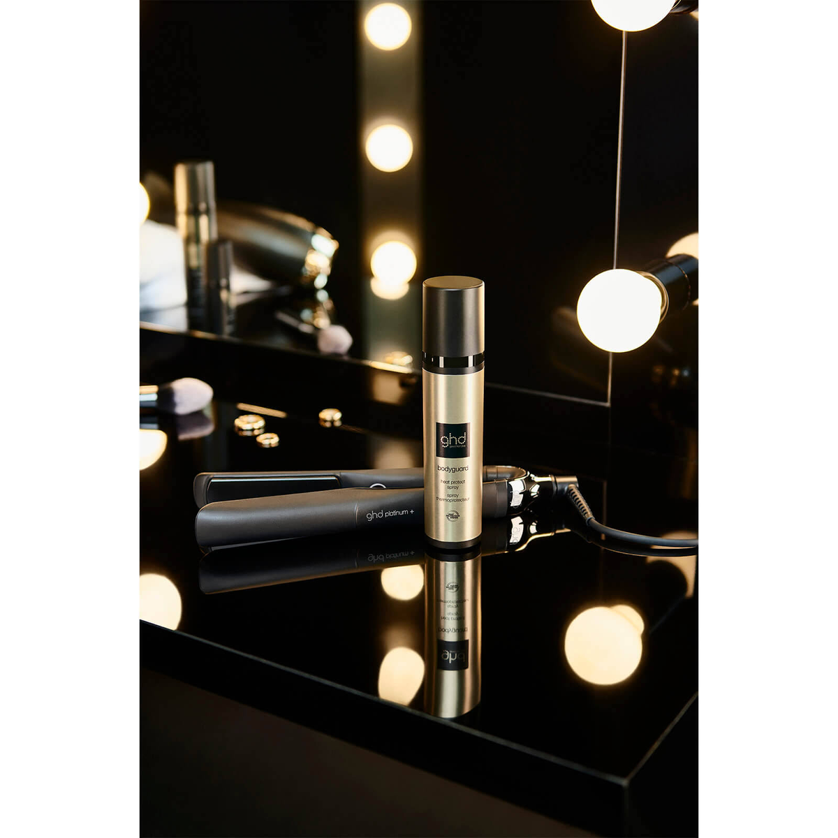 Una bomboletta spray oro e nera e una piastra per capelli su una superficie nera con luci e uno specchio.