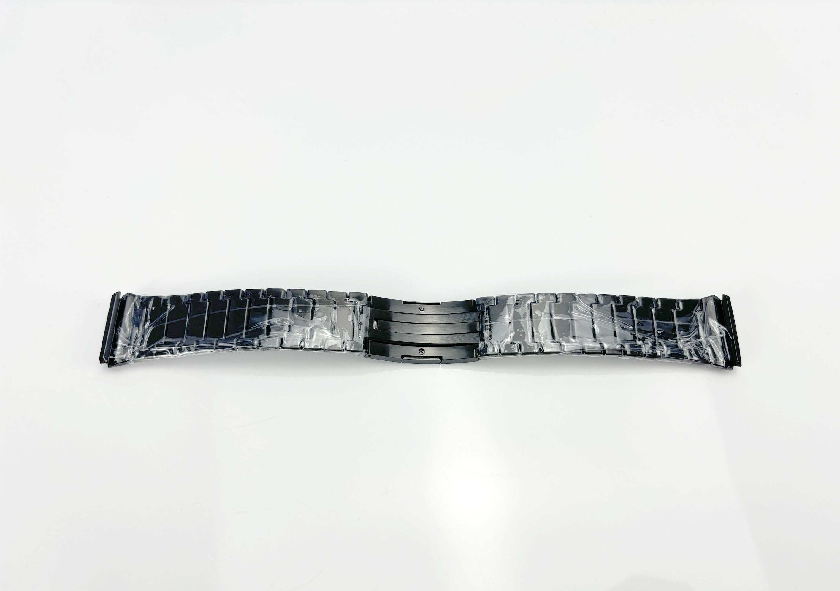 Ein schwarzes Uhrenarmband, zur Hälfte gefaltet, in Klarsichtfolie verpackt, auf weißer Oberfläche liegend.