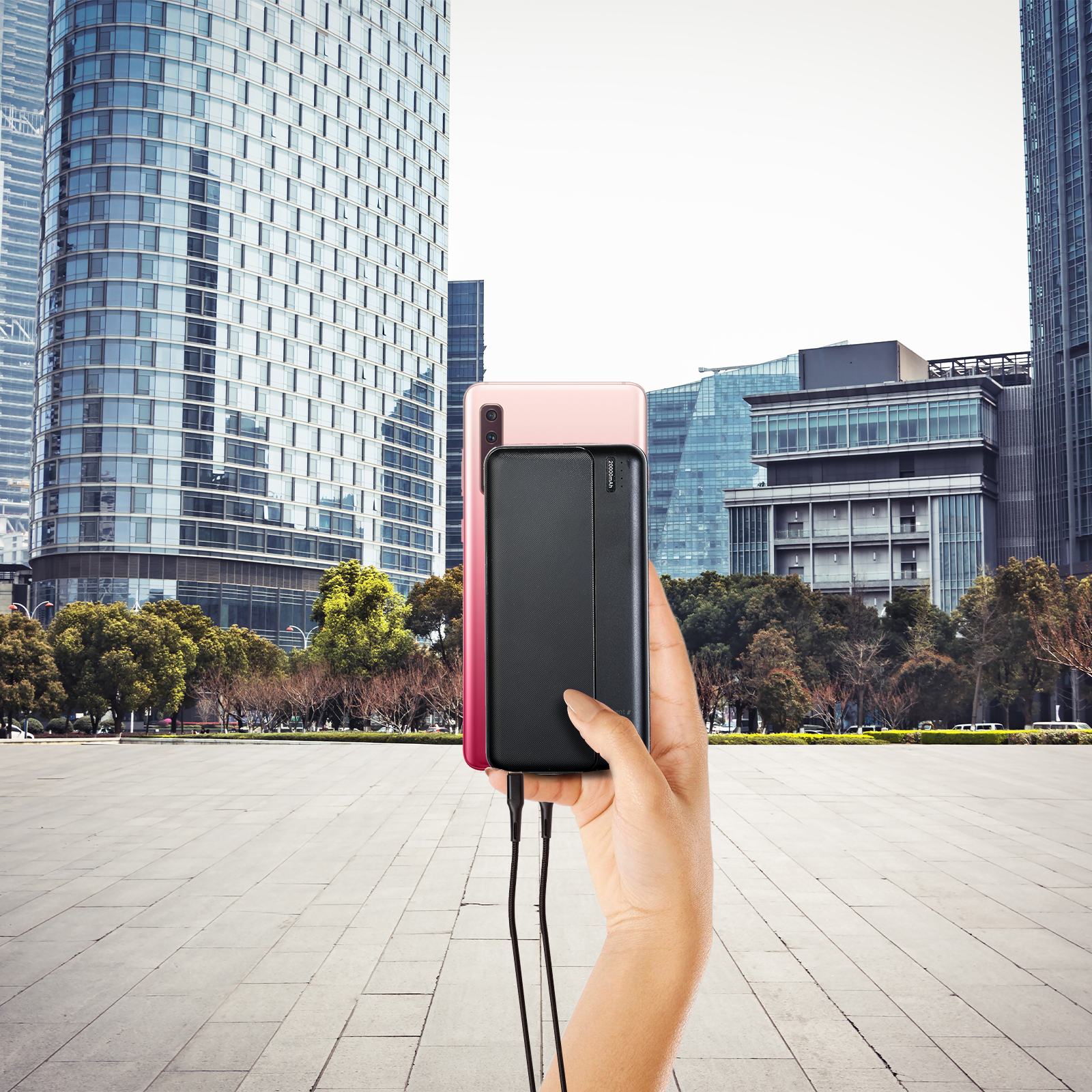 Una mano sosteniendo un power bank negro cargando un teléfono inteligente rosa, con edificios de la ciudad al fondo.
