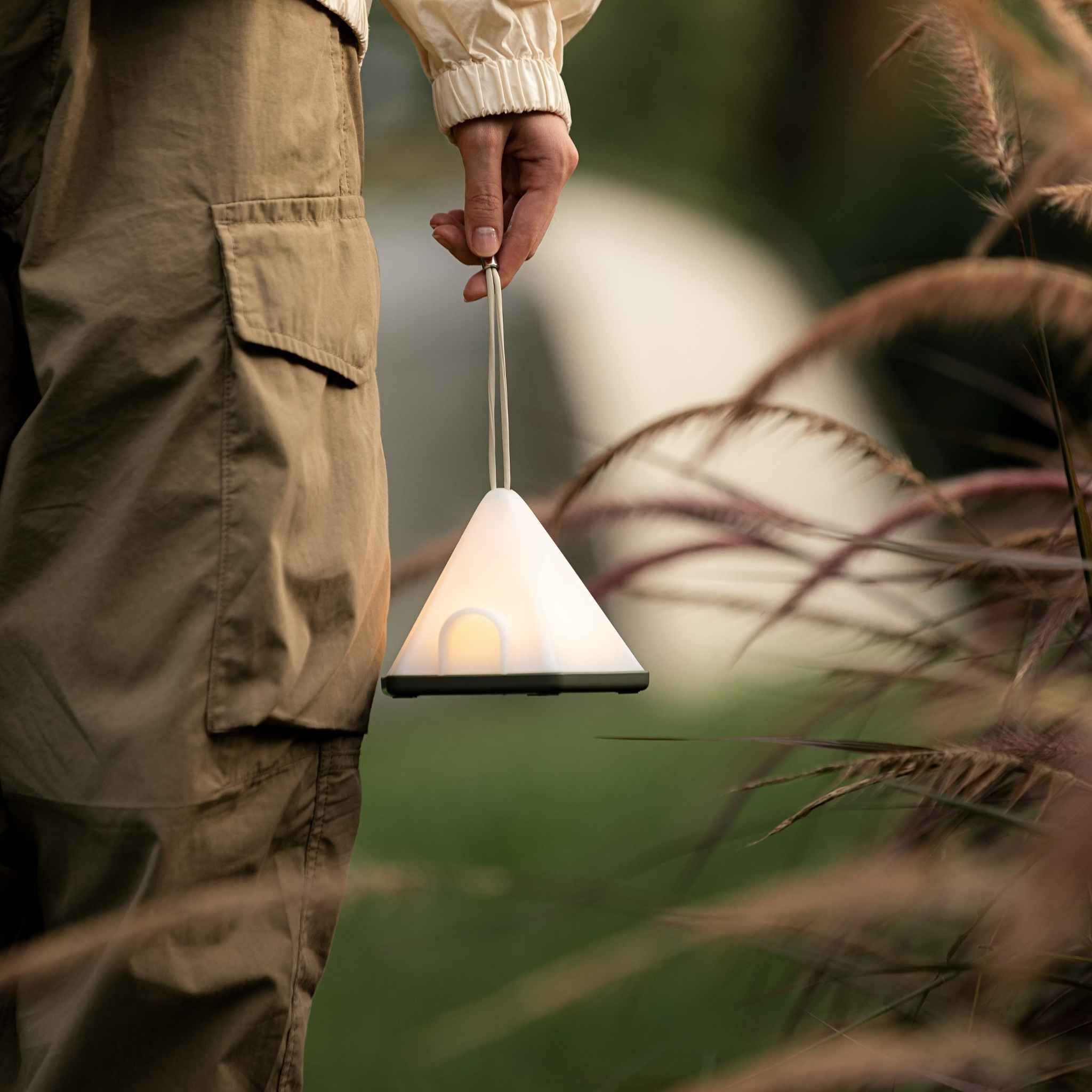 Eine Hand hält eine weiße, dreieckige Lampe mit beigem Sockel, die an einer Schnur hängt, im Freien.